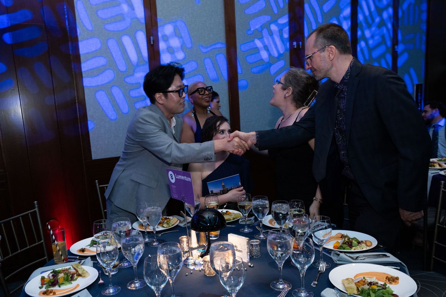 Two guests shake hands across a formal dinner table set with wine glasses and plated meals at a gala.