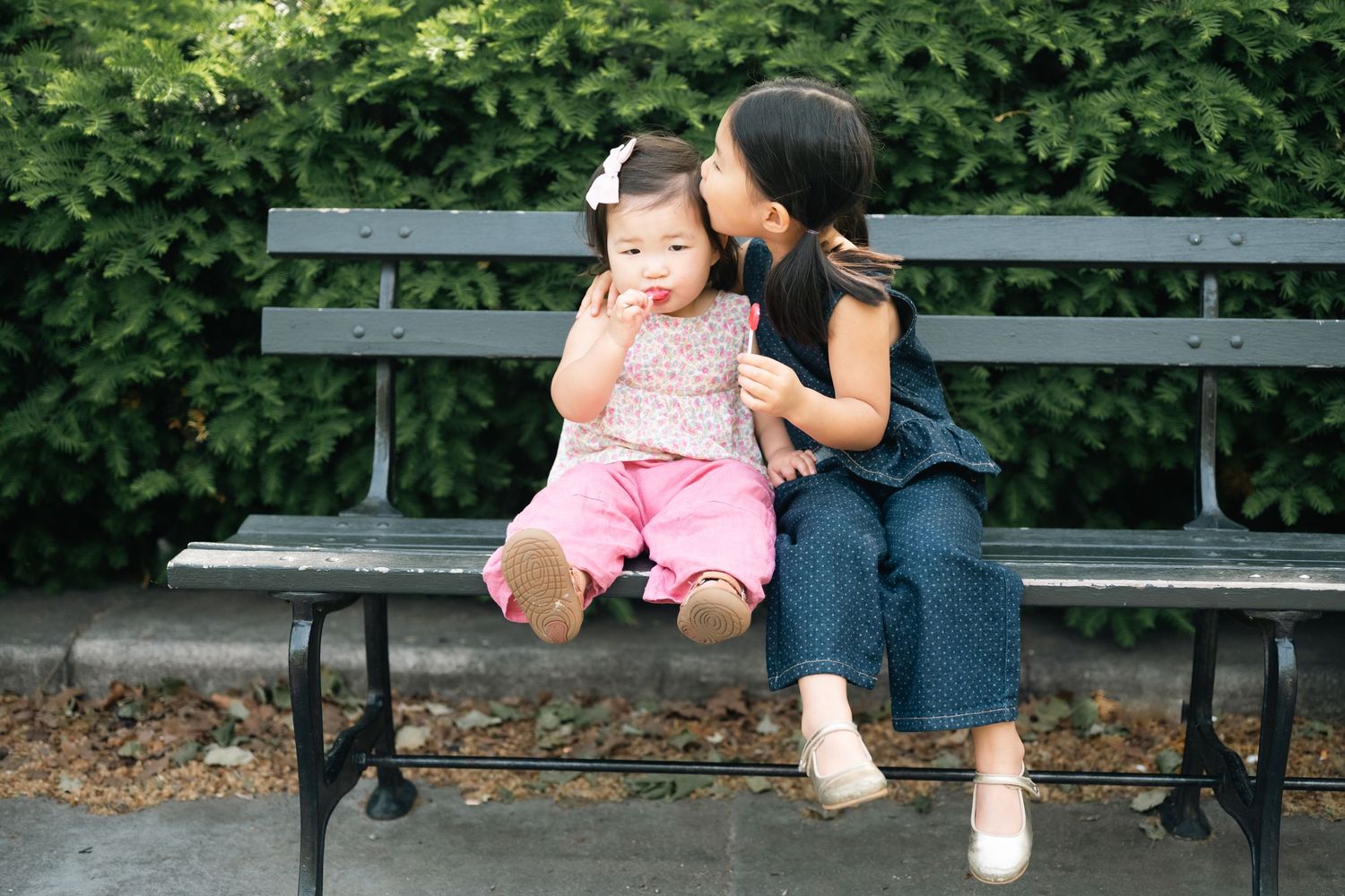 Two young Asian sisters sit on a park bench, one kissing the other's cheek while she eats a snack.