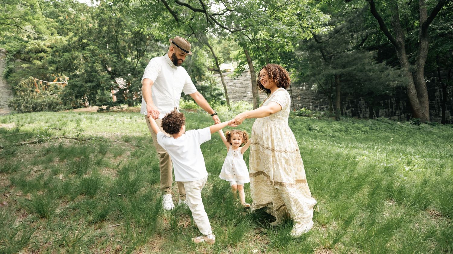 A joyful family of four plays together in a lush green park, parents swinging their children outdoors.