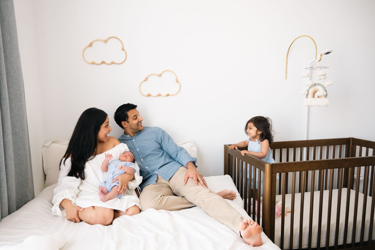 Family lounging on bed with cloud wall decor and wooden crib in background.