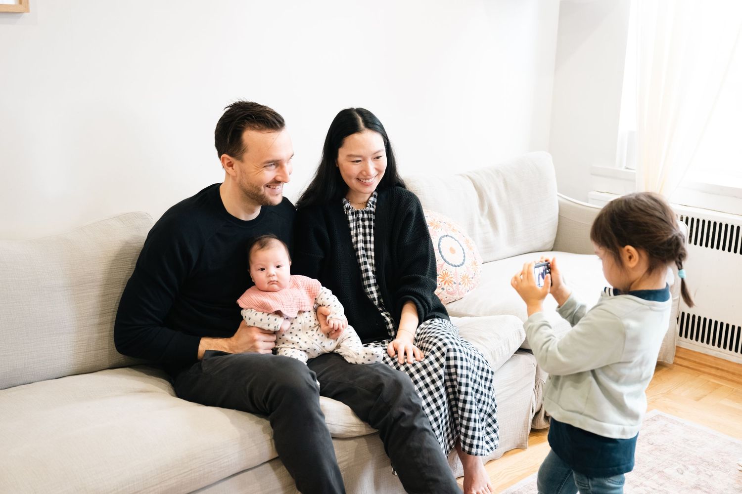 Family members sit together on a cozy white couch in a bright living room during a casual photo session.