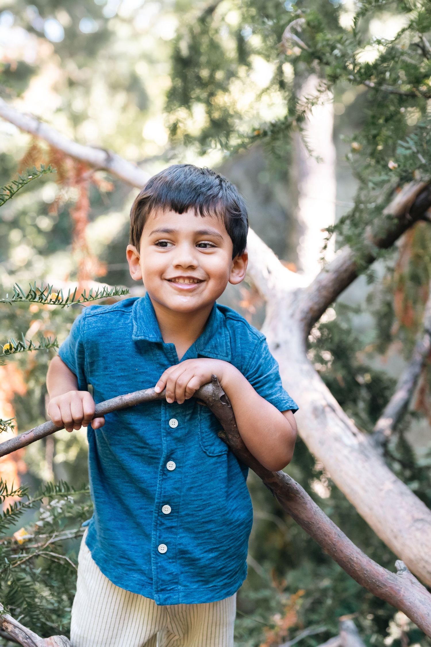 A young child in a blue button-up shirt plays outdoors among trees and foliage with a joyful expression.