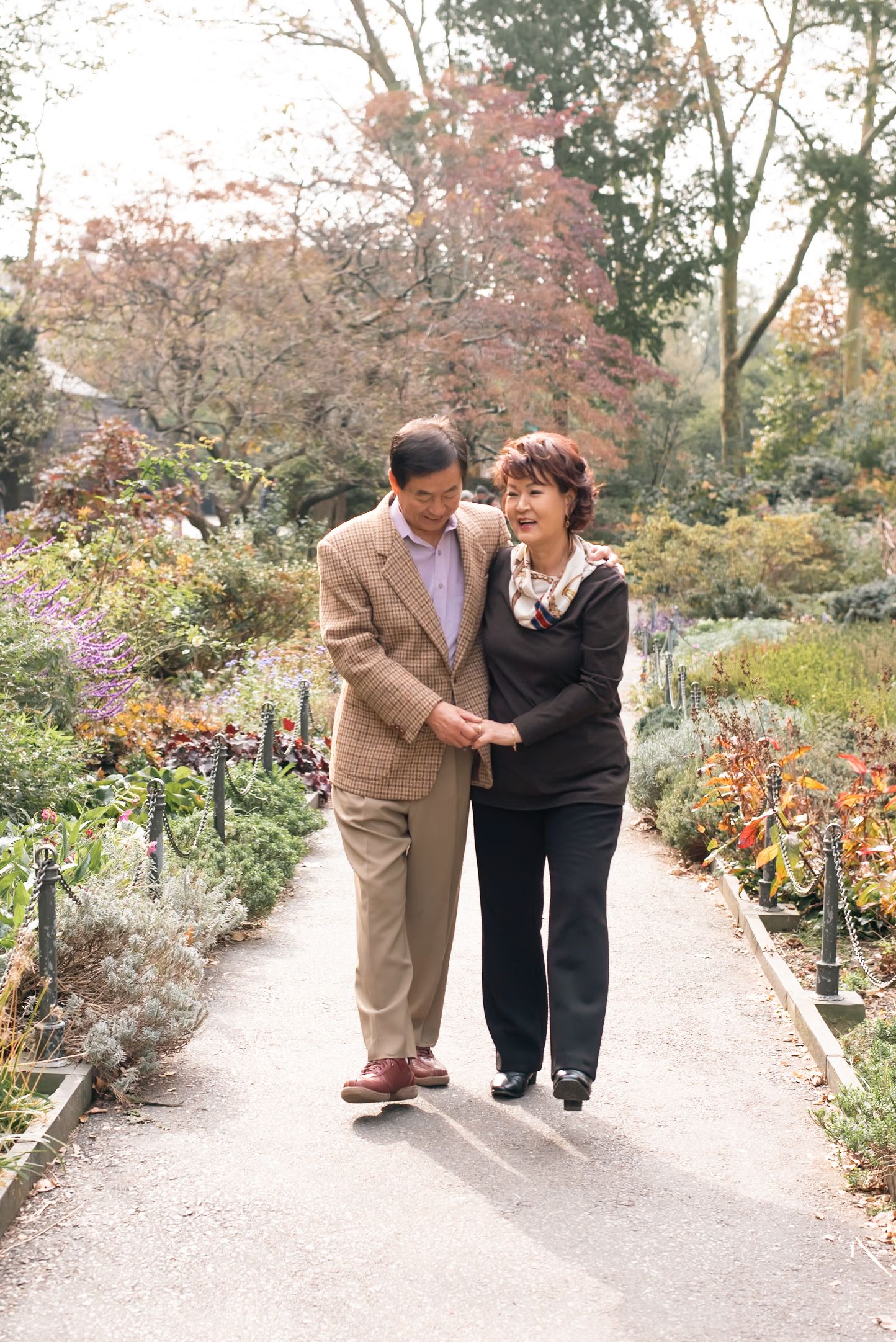 Two people walk together along a garden path surrounded by colorful autumn foliage.
