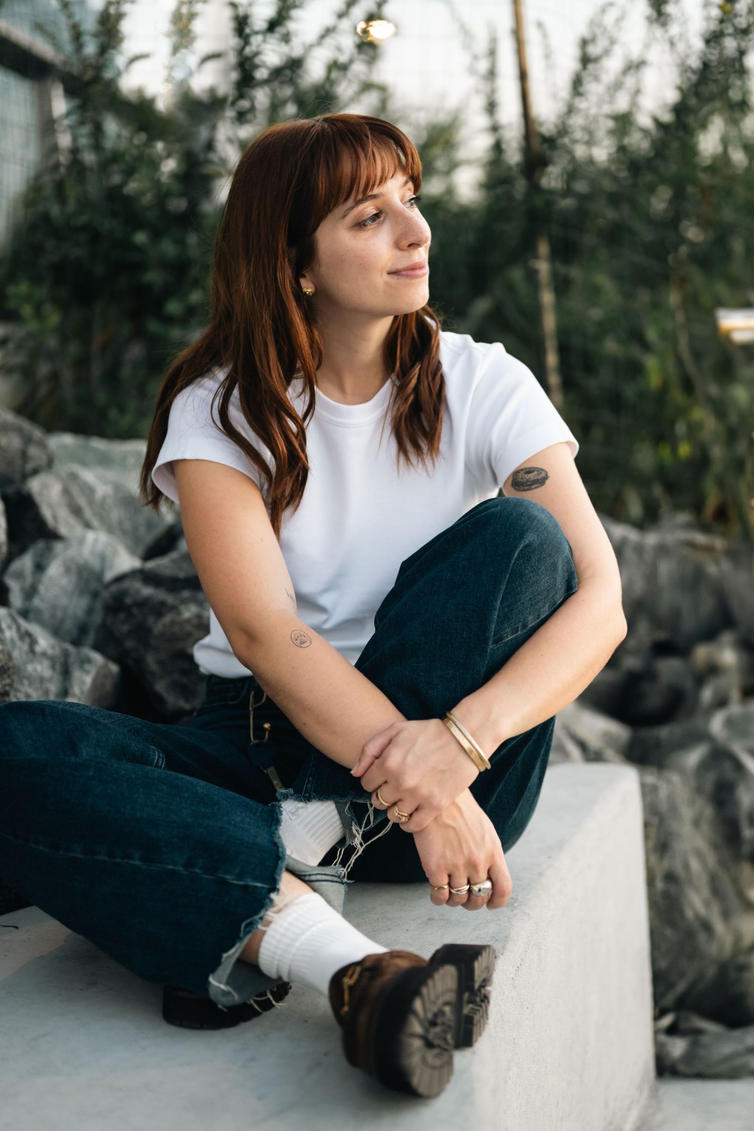 Casual outdoor photoshoot featuring someone in white t-shirt and jeans sitting on concrete near rocks.