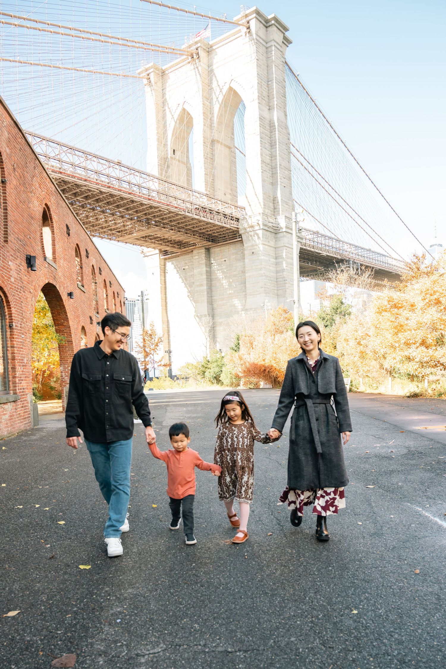 A group of people walking together on a path beneath the Brooklyn Bridge on a sunny autumn day.