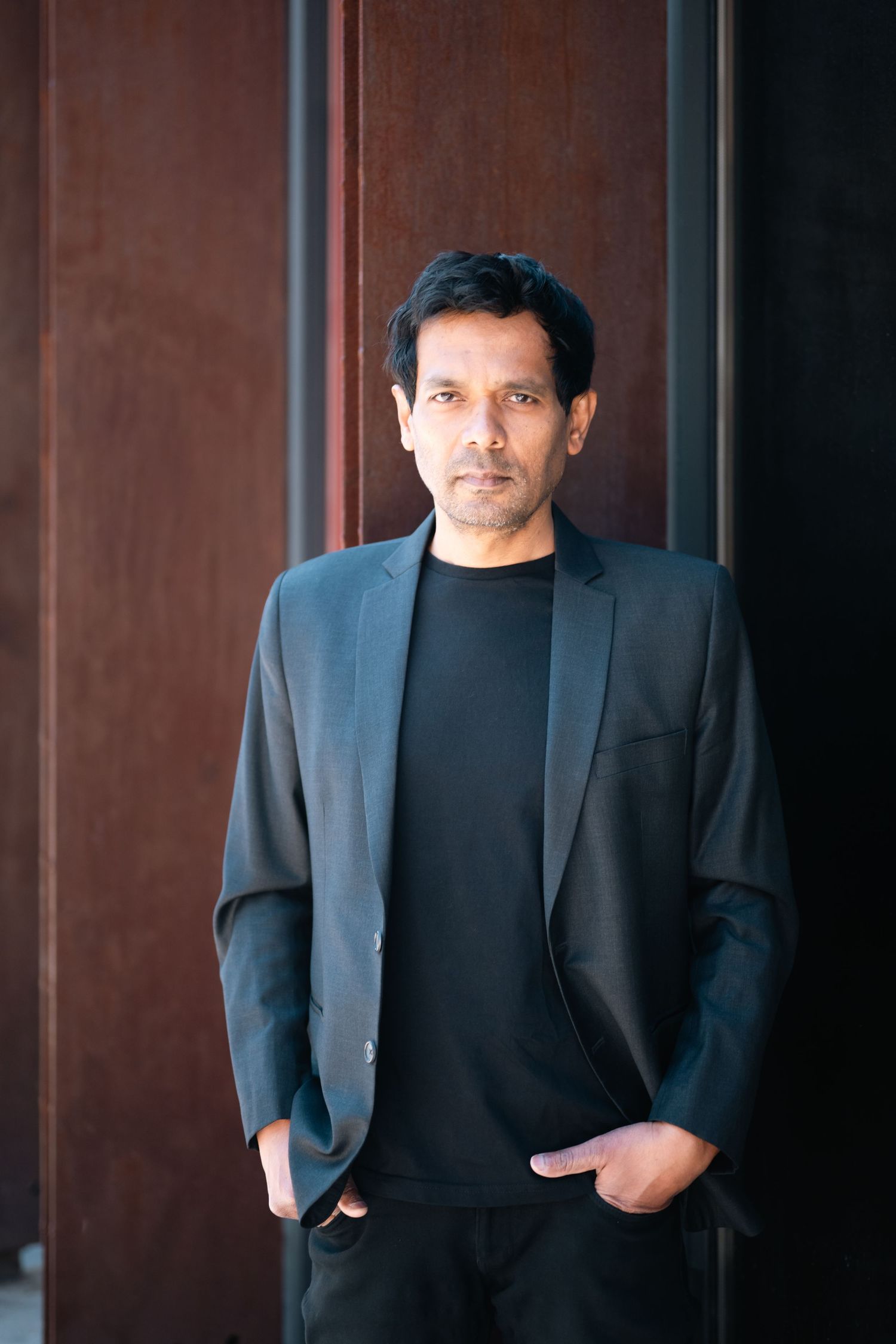 A confident man in a black blazer and t-shirt stands with hands in pockets against a dark wooden wall.