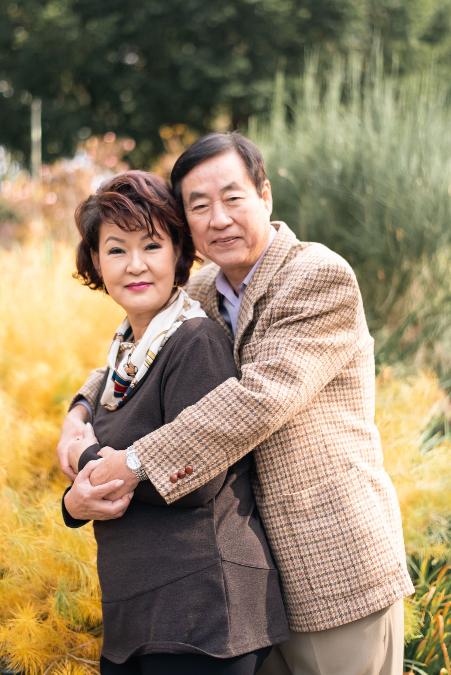 Couple embracing outdoors with autumn foliage in background, wearing casual attire.
