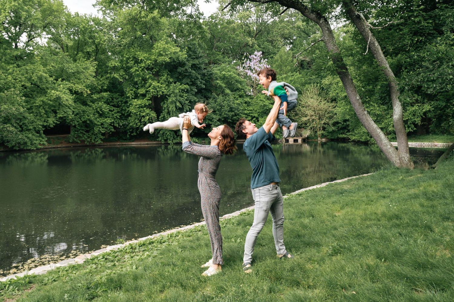 Family playing outdoors near a pond, parents lifting two young children in a lush green park setting.