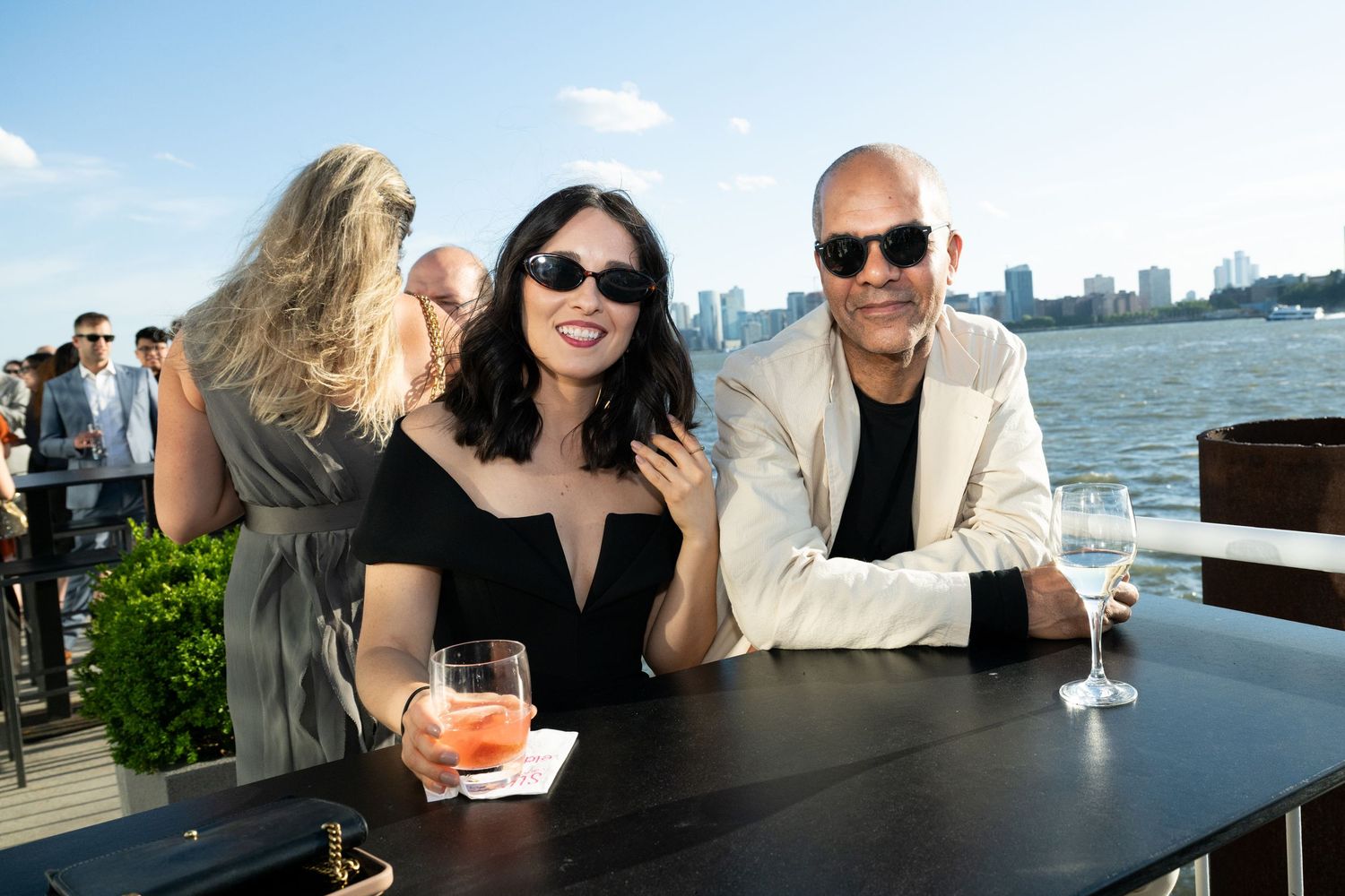 A couple smiles at an outdoor waterfront event, woman holding a cocktail, city skyline visible in background.