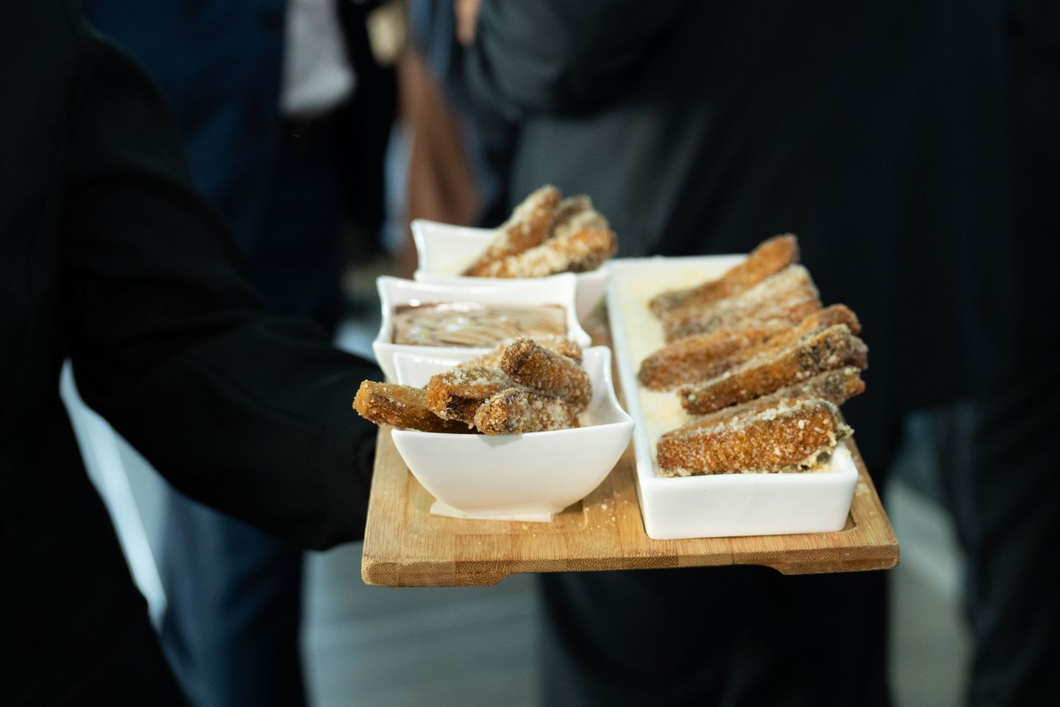 A server holds a wooden tray with white bowls filled with assorted breaded appetizers at a catered event.