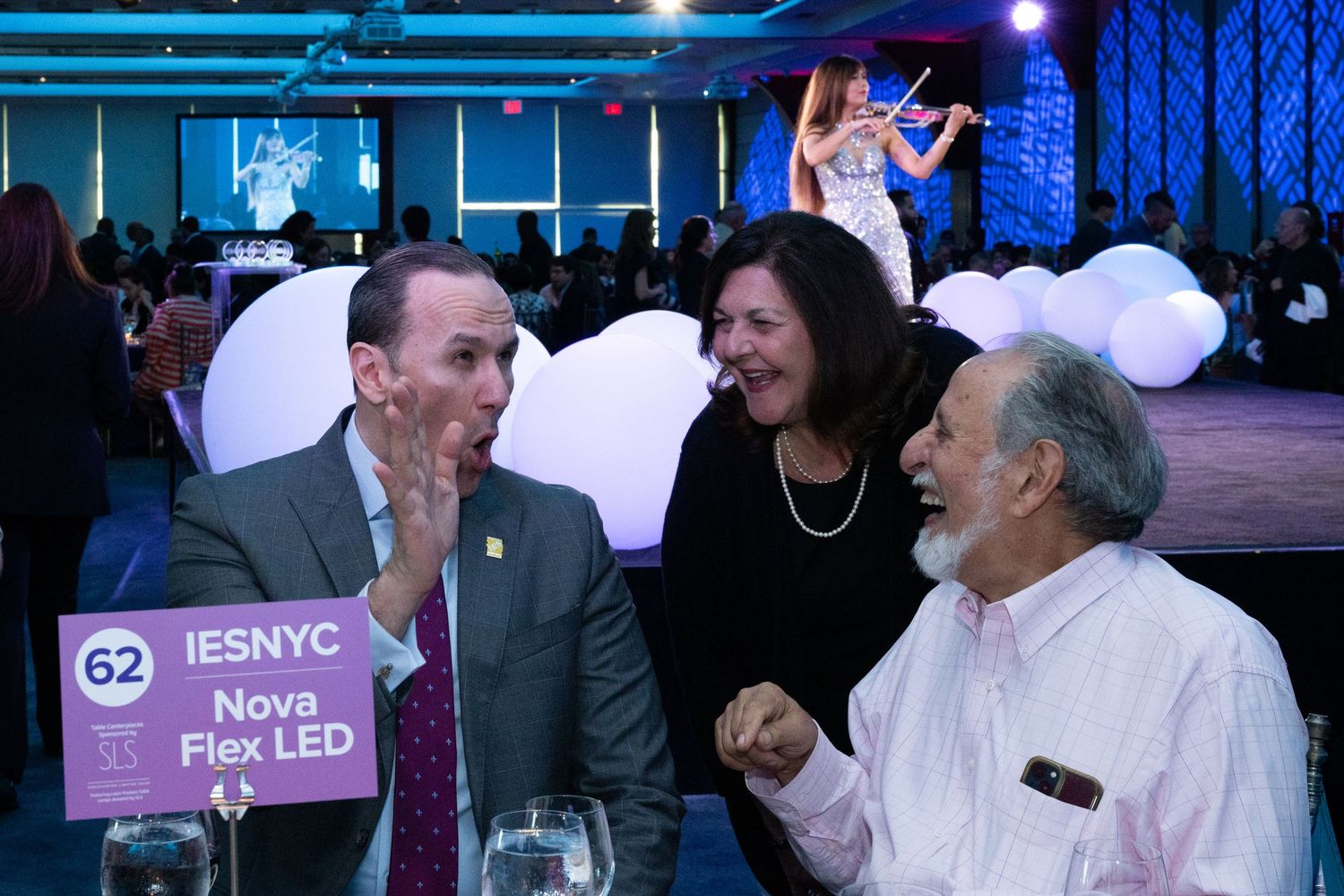 Three guests laugh and converse at table 62 during a formal gala dinner with live violin performance.