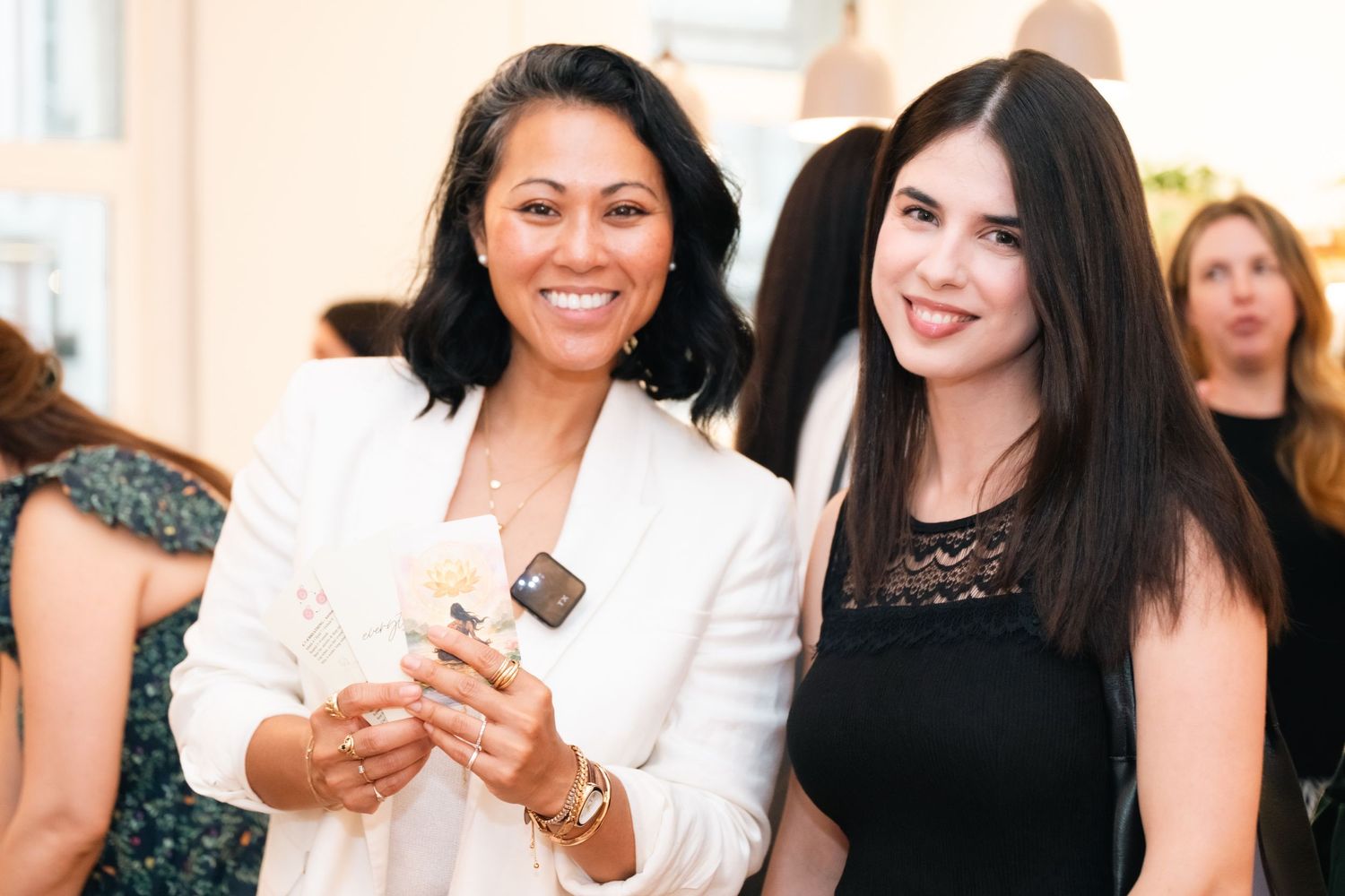 Two smiling women pose together at an elegant event, one in a white blazer holding a phone, the other in a black top.