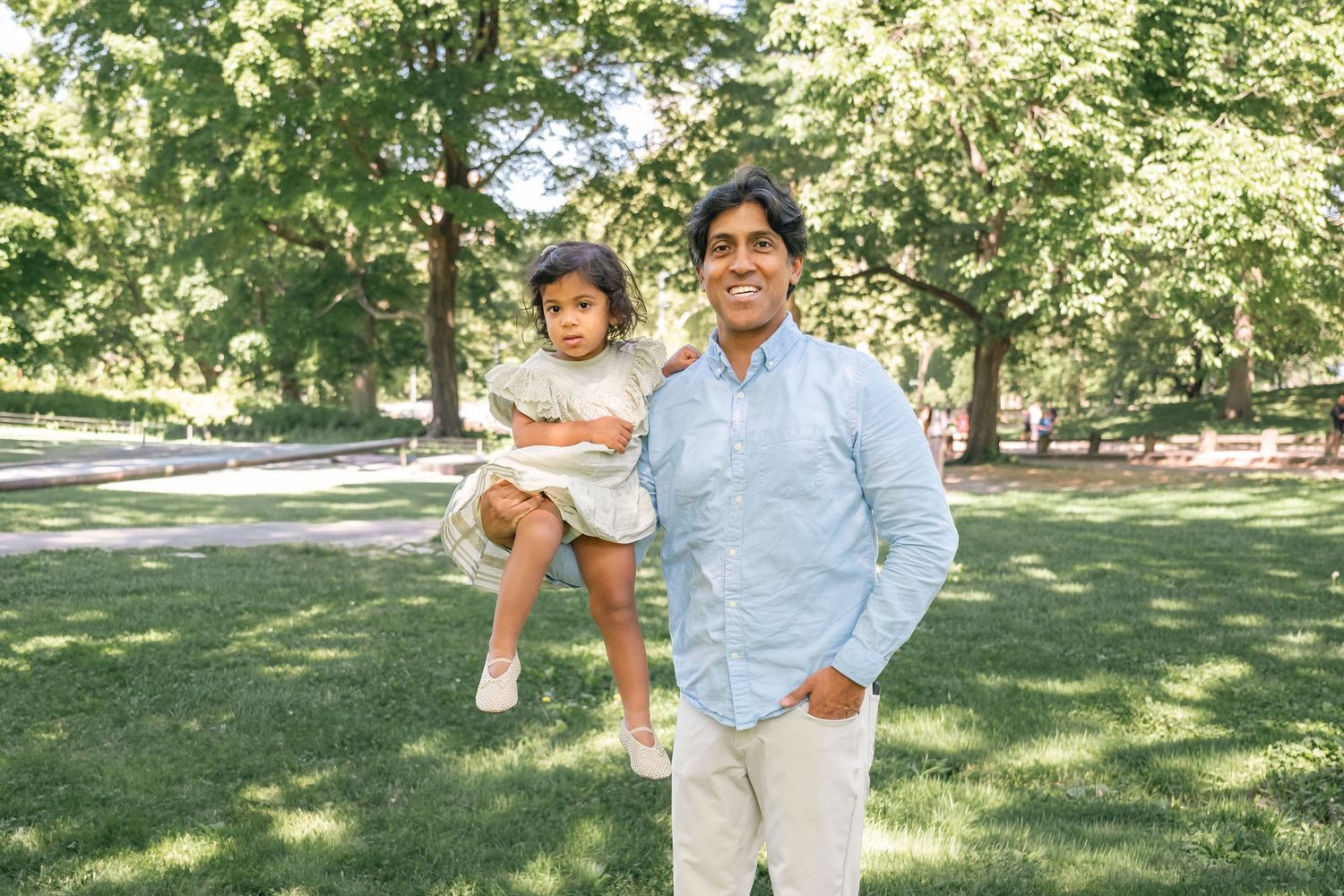 A man holds a young child in a sunny green park, both smiling outdoors among lush trees.