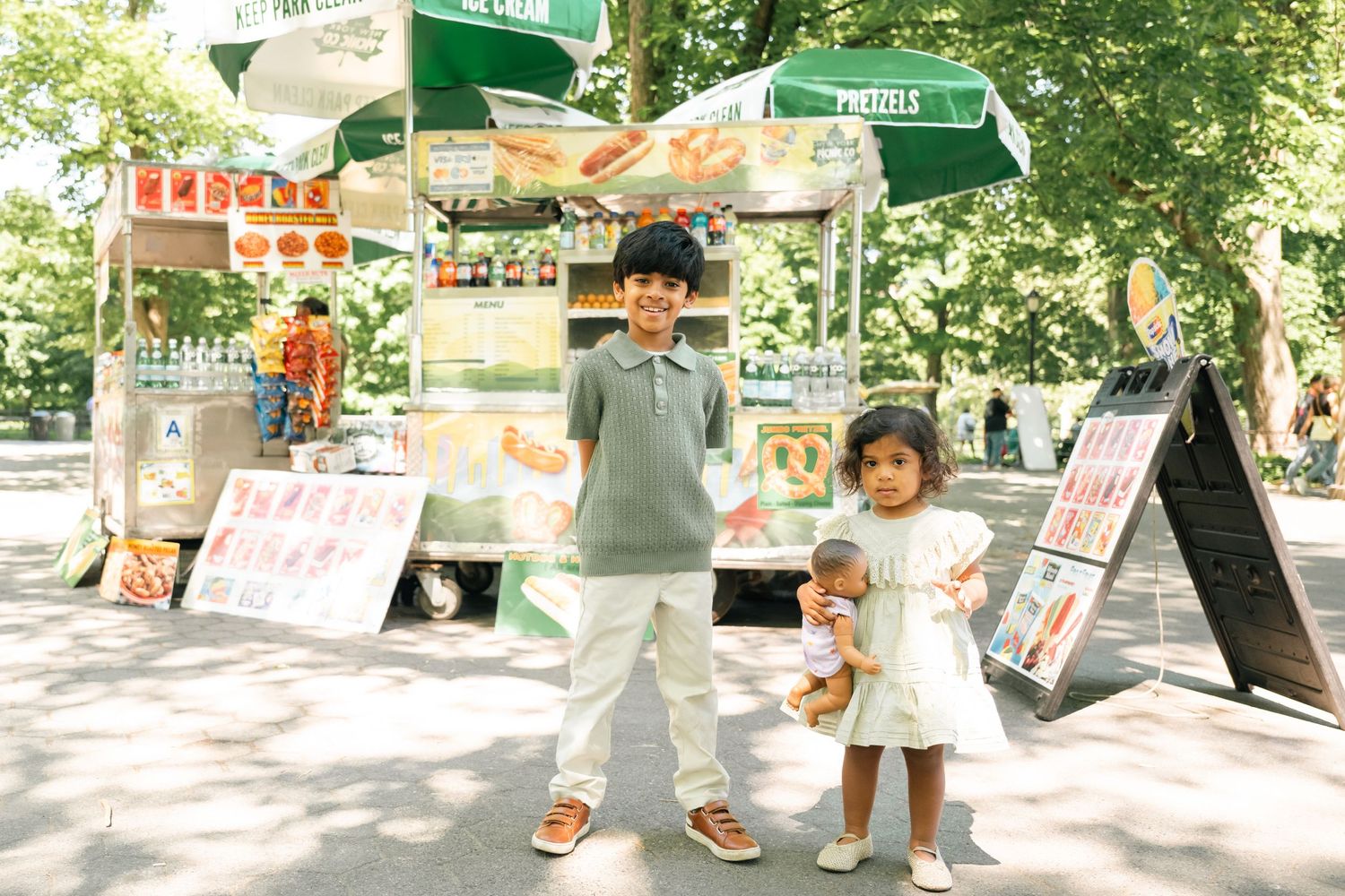 Two children stand smiling in front of a food vendor cart in a sunny park, with a boy and girl posing happily.