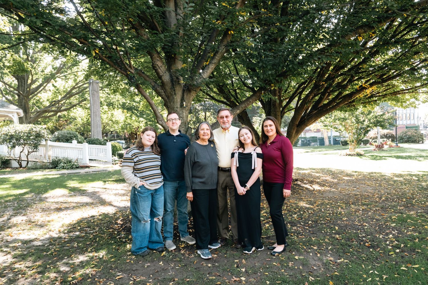 A group poses for a casual photo under large shade trees in a park setting.