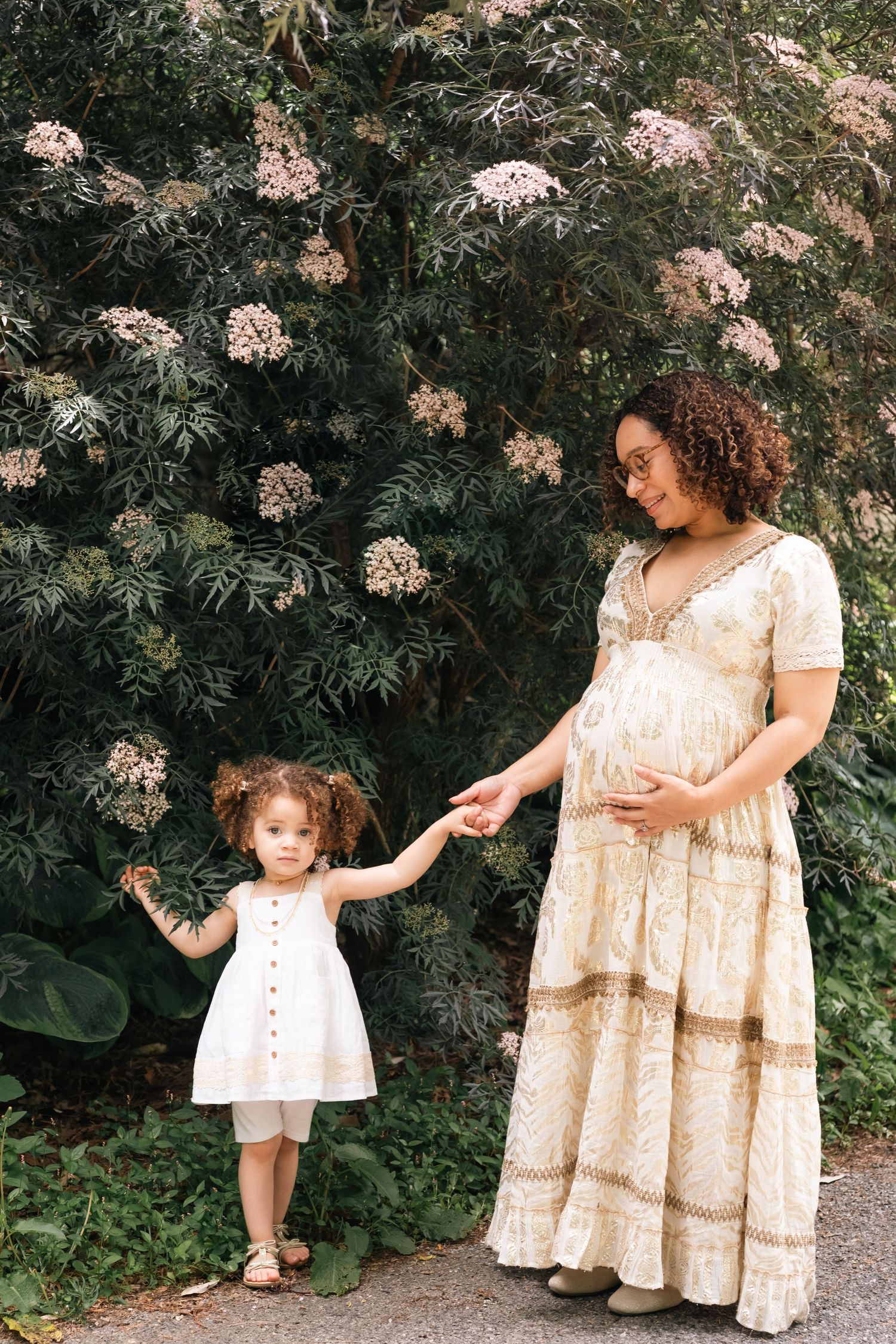 Pregnant woman in floral dress holds hands with toddler girl in white dress beside blooming green bushes outdoors.