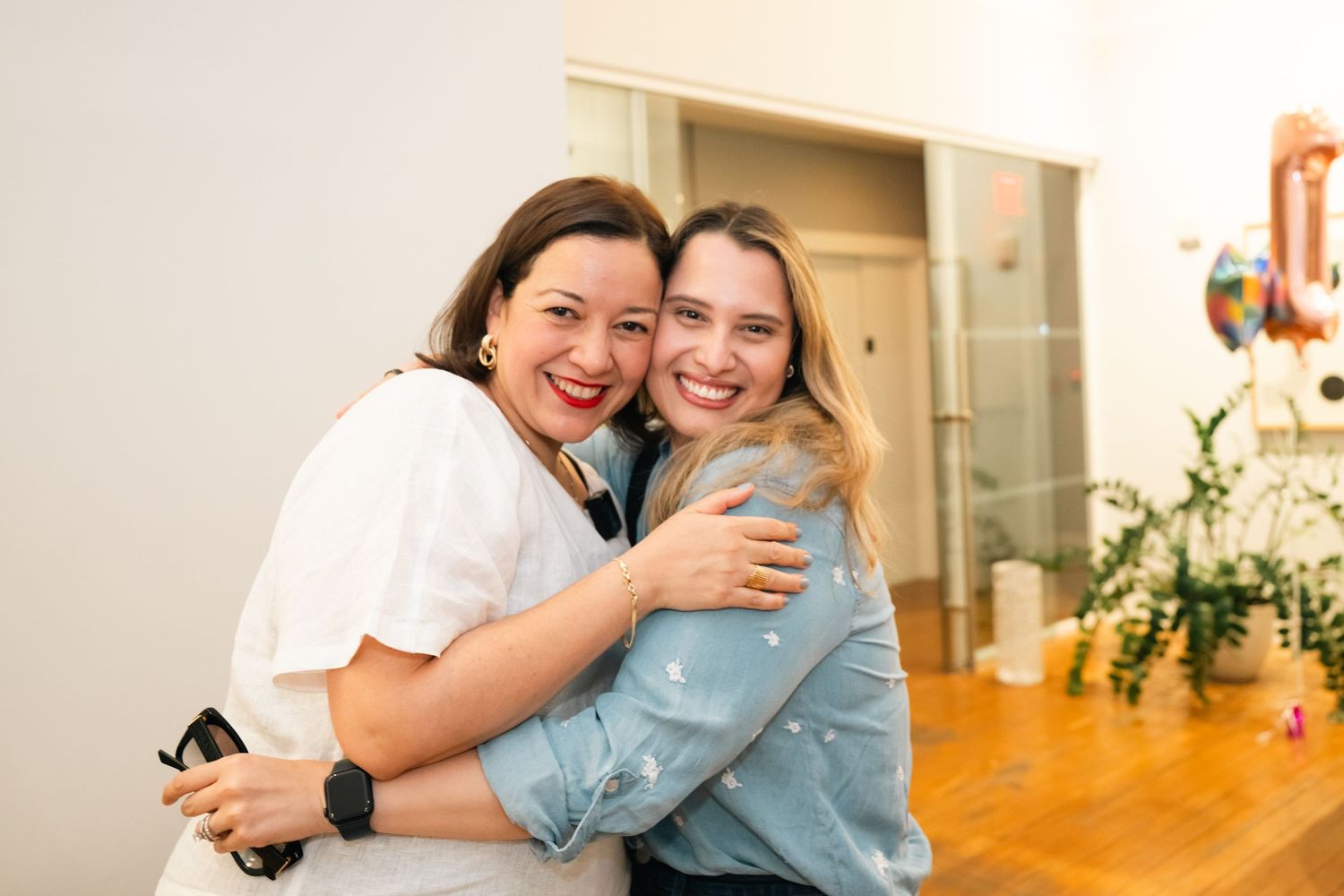 Two women smiling and hugging each other warmly indoors at a casual gathering.