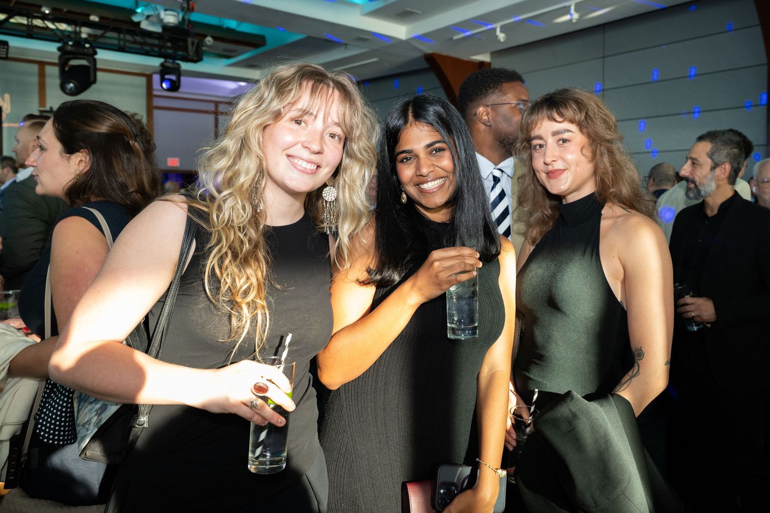 Three young women smile and hold drinks together at an elegant evening party event.