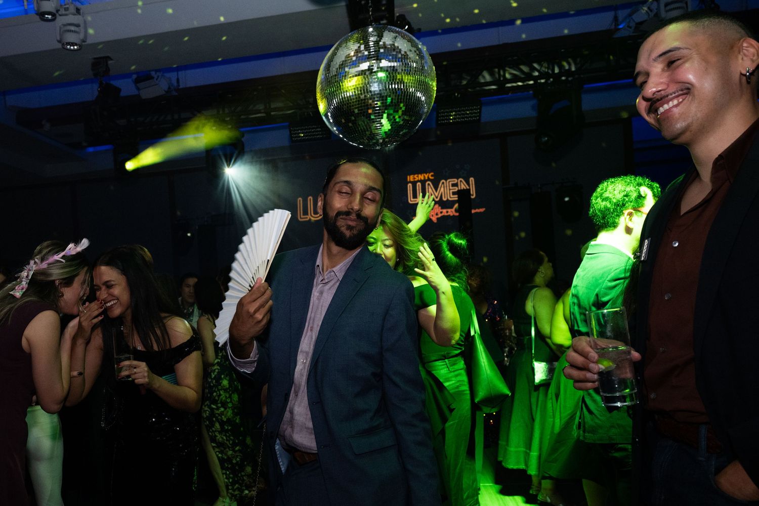 A man dances joyfully holding a white fan under a disco ball at a vibrant gala party.