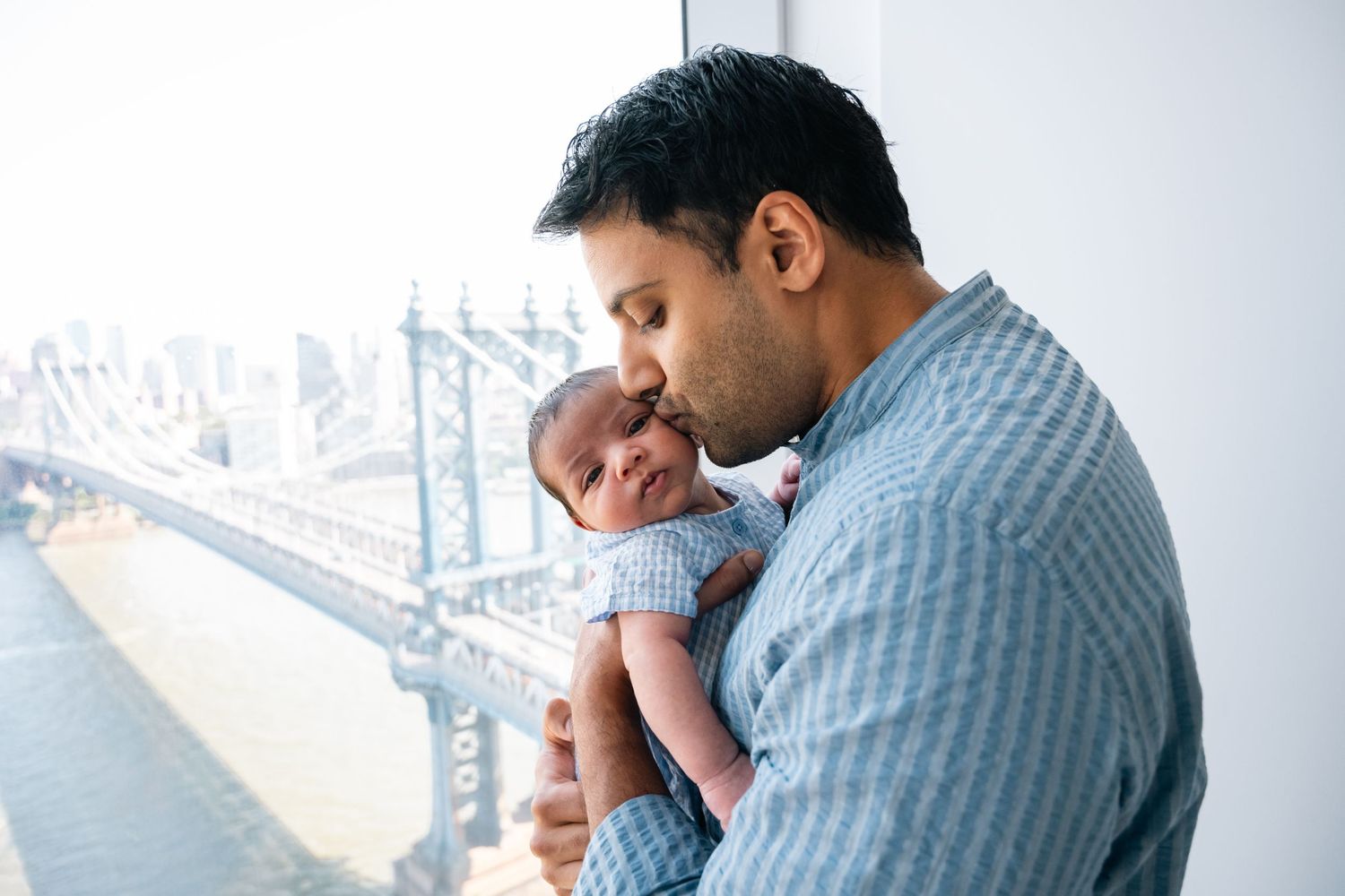 Parent cradling and kissing newborn baby near a window overlooking a city bridge.
