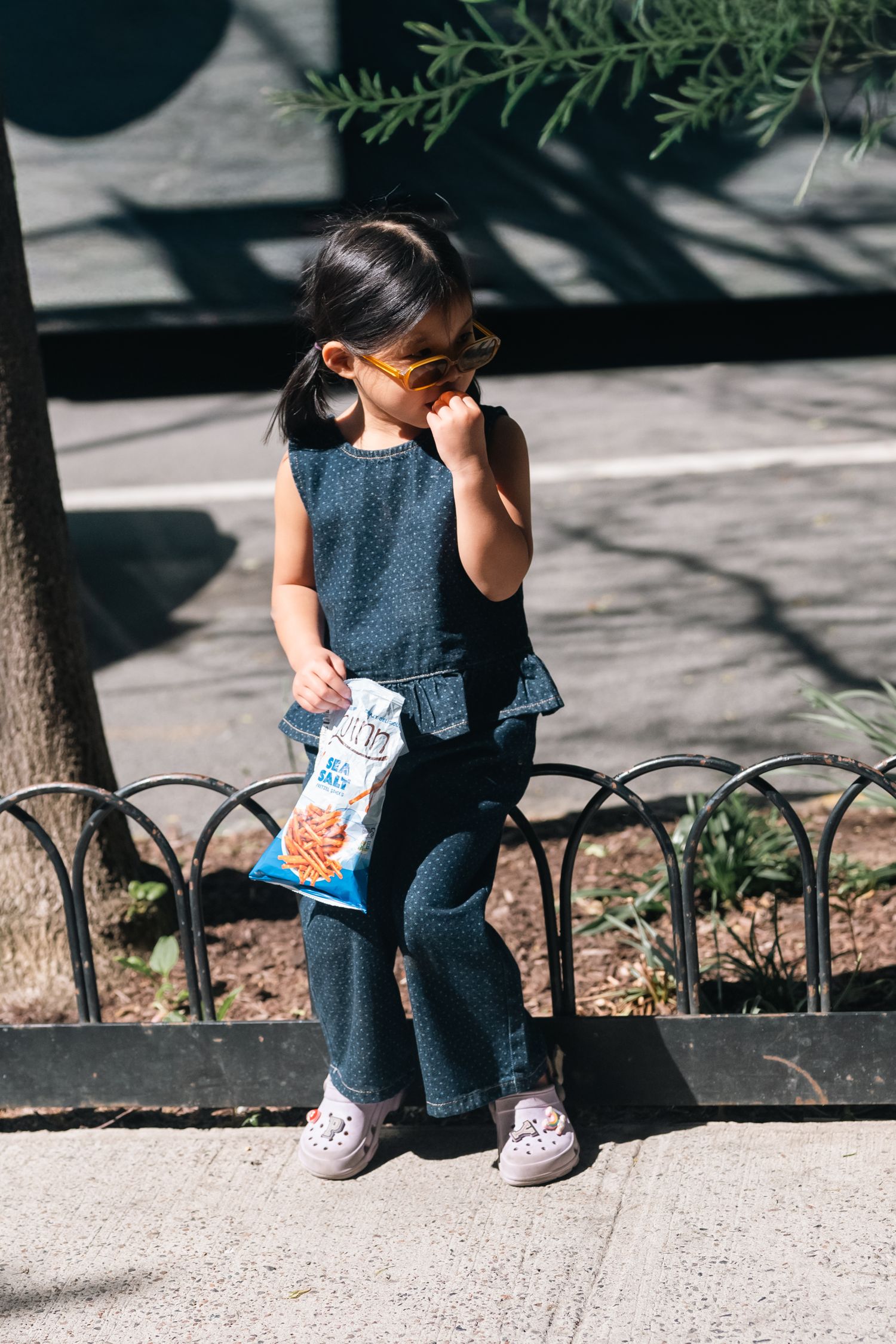 Young girl in denim jumpsuit wearing orange sunglasses holding a snack bag while sitting on a curb outdoors.