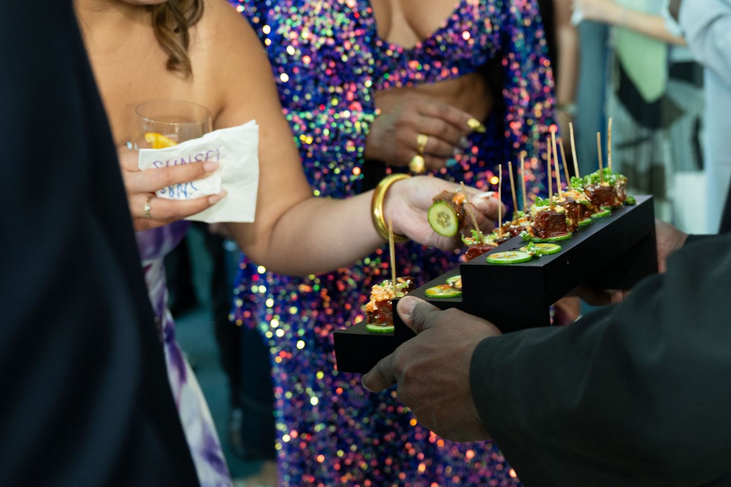 Guests in sequin dresses selecting elegant appetizers from a black tray at a glamorous catered event.