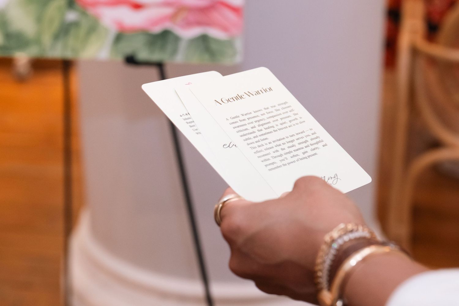A hand holding white cards with text, adorned with gold rings and bracelets, at an elegant event.