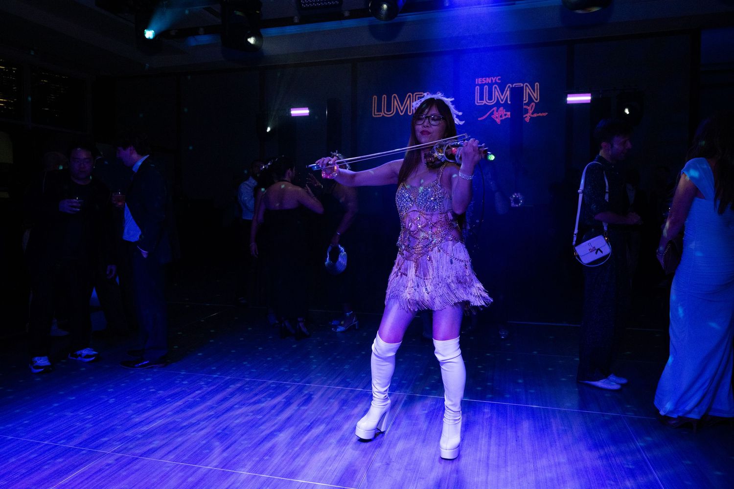 A performer in a sparkly dress and white boots dances on a blue-lit stage.