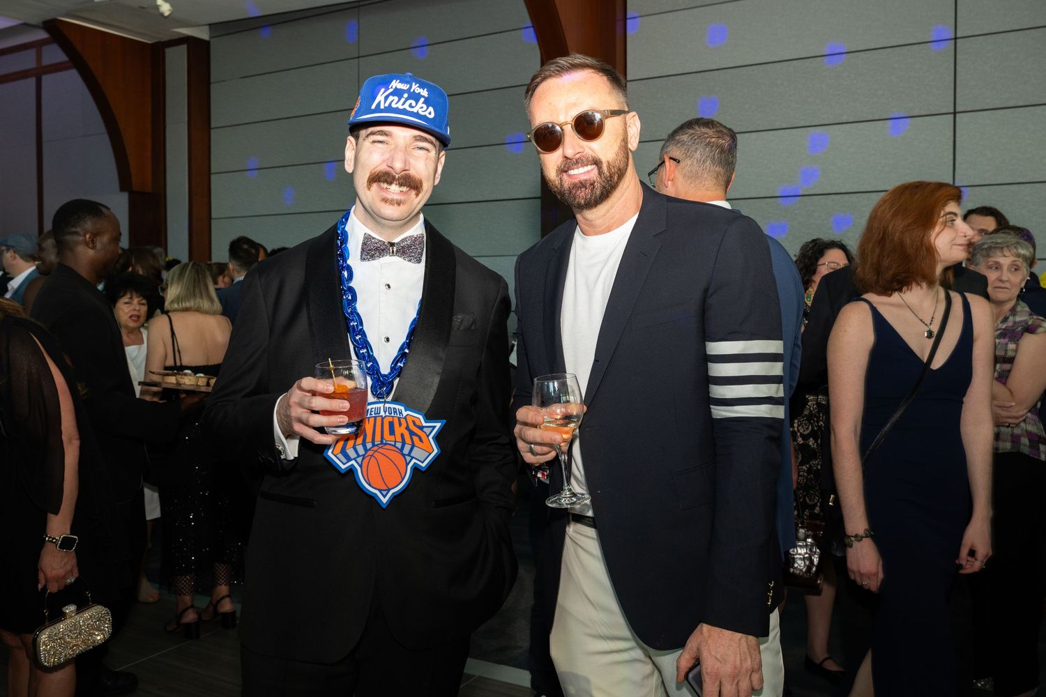 Two men posing at an indoor gala, one wearing a Knicks hat and sports jersey, holding wine glasses.