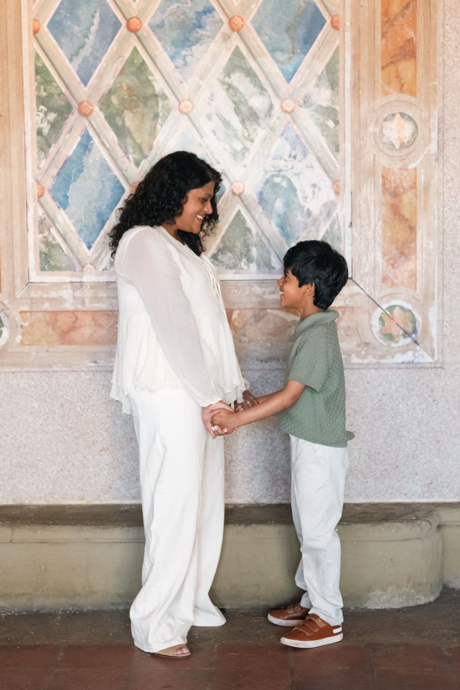 A pregnant woman in white holds hands with a young boy in a green shirt, sharing a tender moment outdoors.