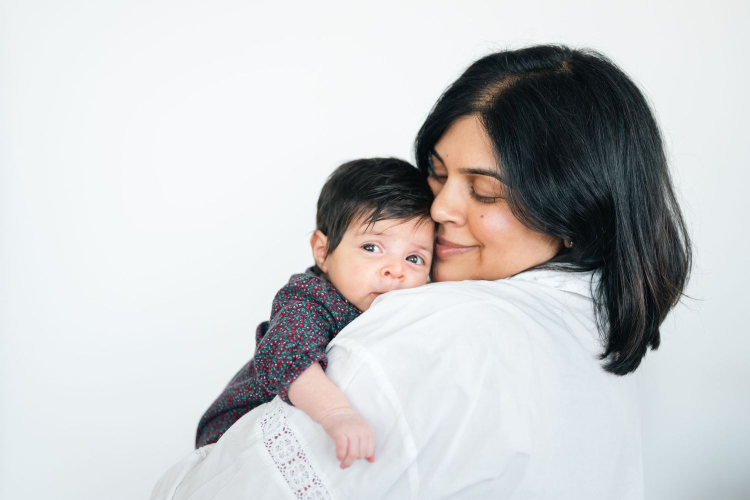 A loving mother with dark hair gently holds her baby close, sharing a tender embrace against a white background.