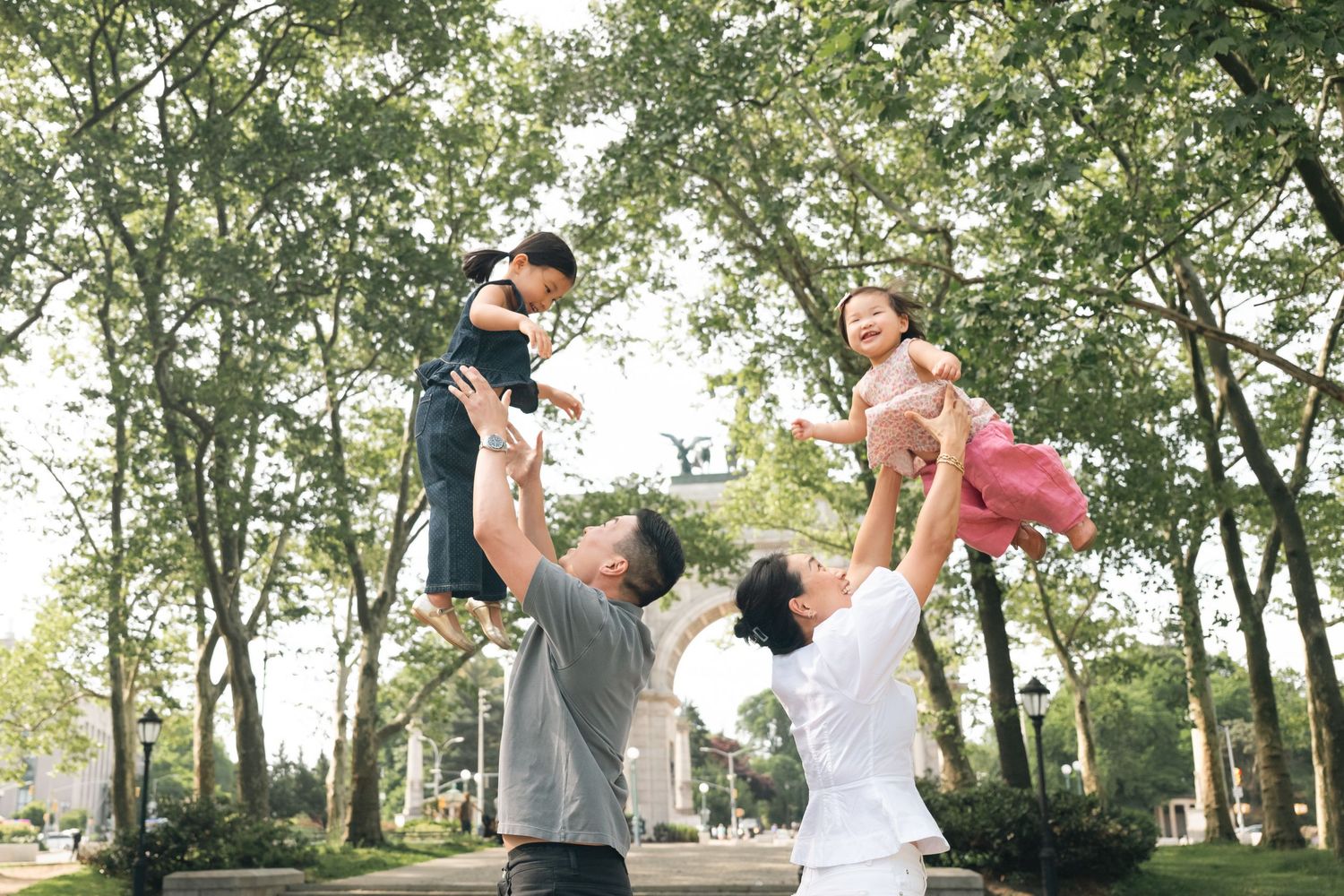 Two parents joyfully toss their young children into the air in a sunny tree-lined park.