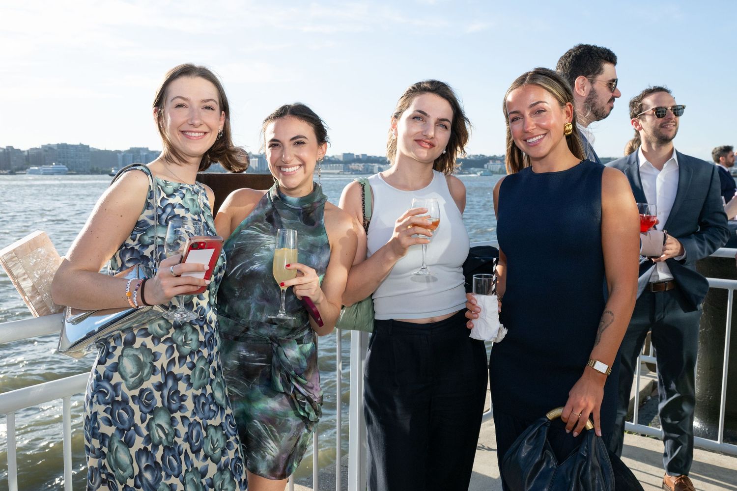 Four women pose together smiling at an outdoor waterfront party on a sunny day holding drinks.