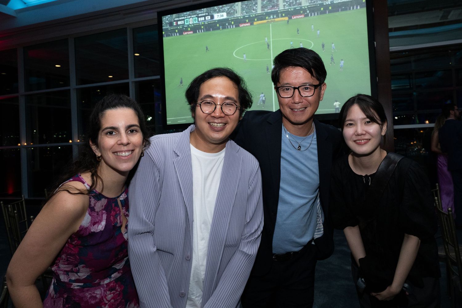 Four colleagues smile together at a networking event with a soccer match playing on a large screen behind them.