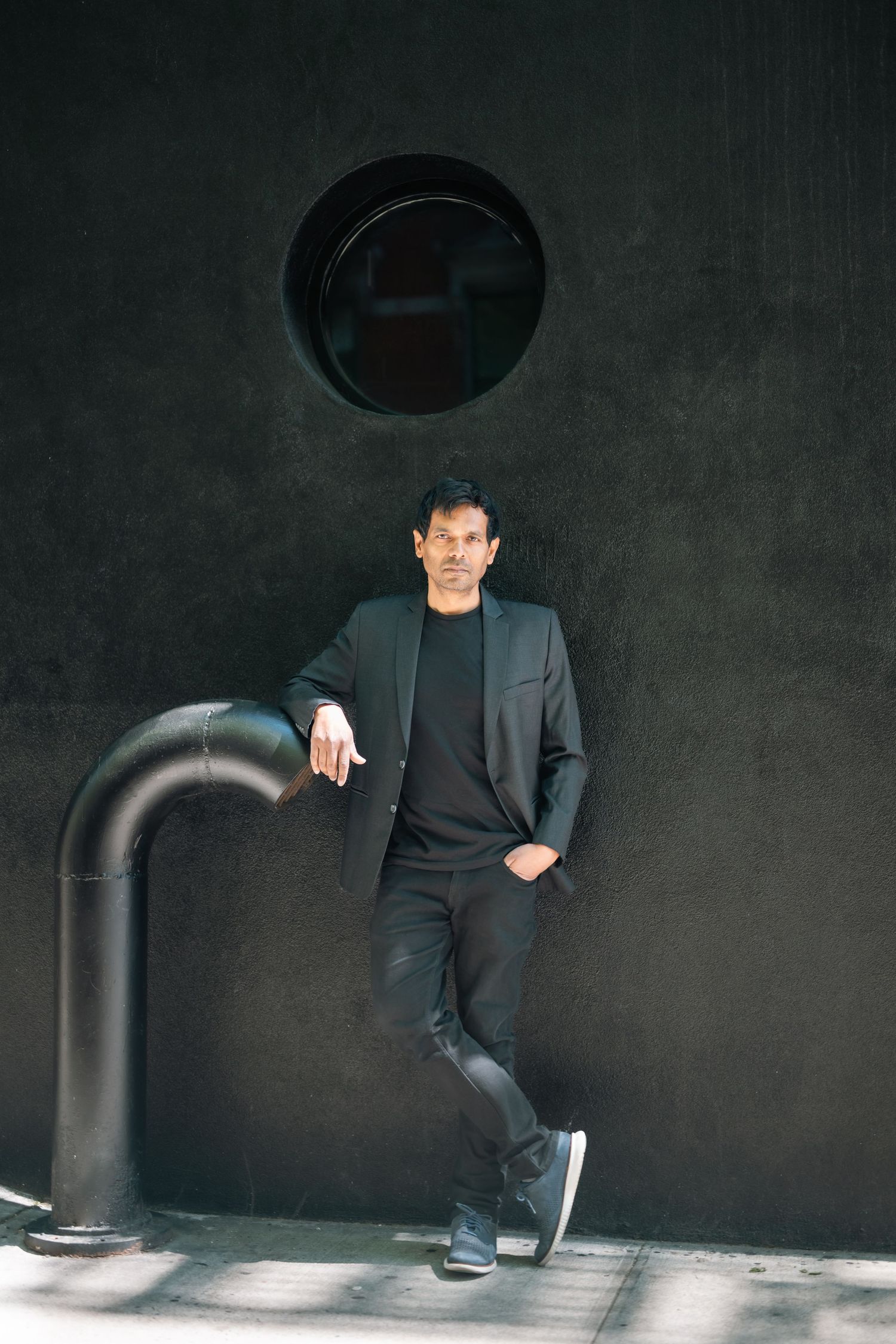 Man in all-black outfit leaning against dark wall with circular window and pipe.