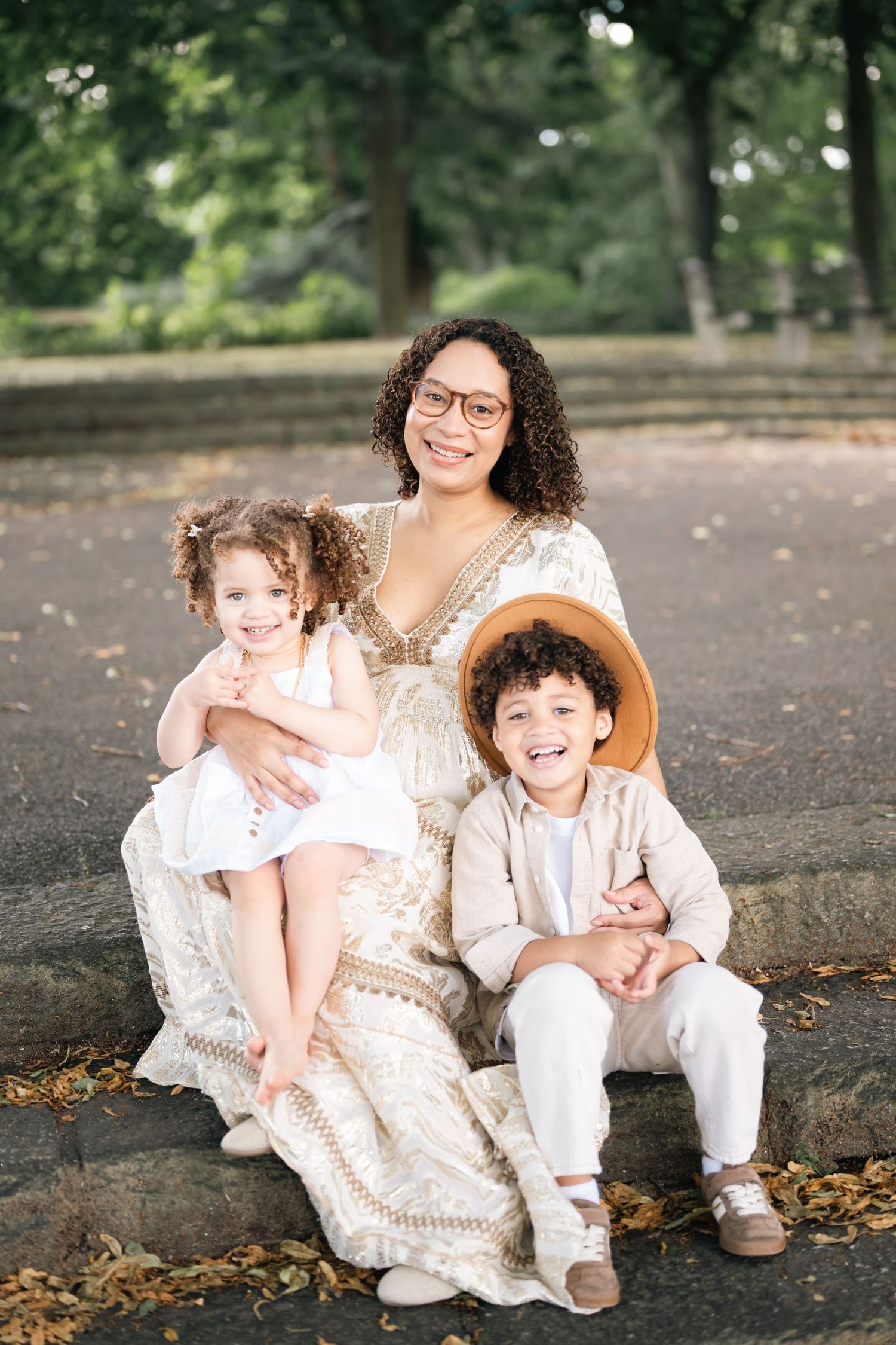 A smiling woman in a floral dress sits outdoors with two young children, a toddler girl and a boy in a straw hat.