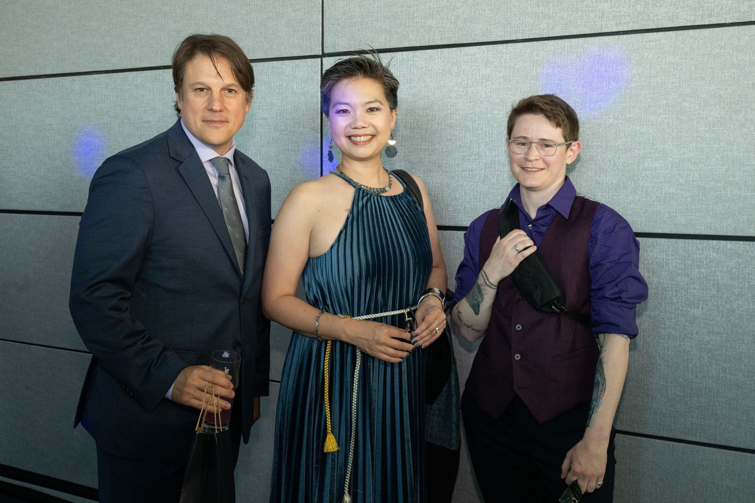 Three elegantly dressed guests pose together smiling at a formal gala event with blue lighting.
