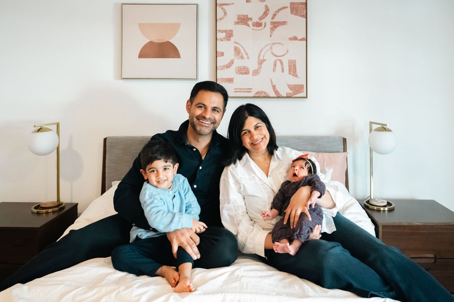Smiling family of four posing on a bed, parents holding two young children in a bright bedroom.