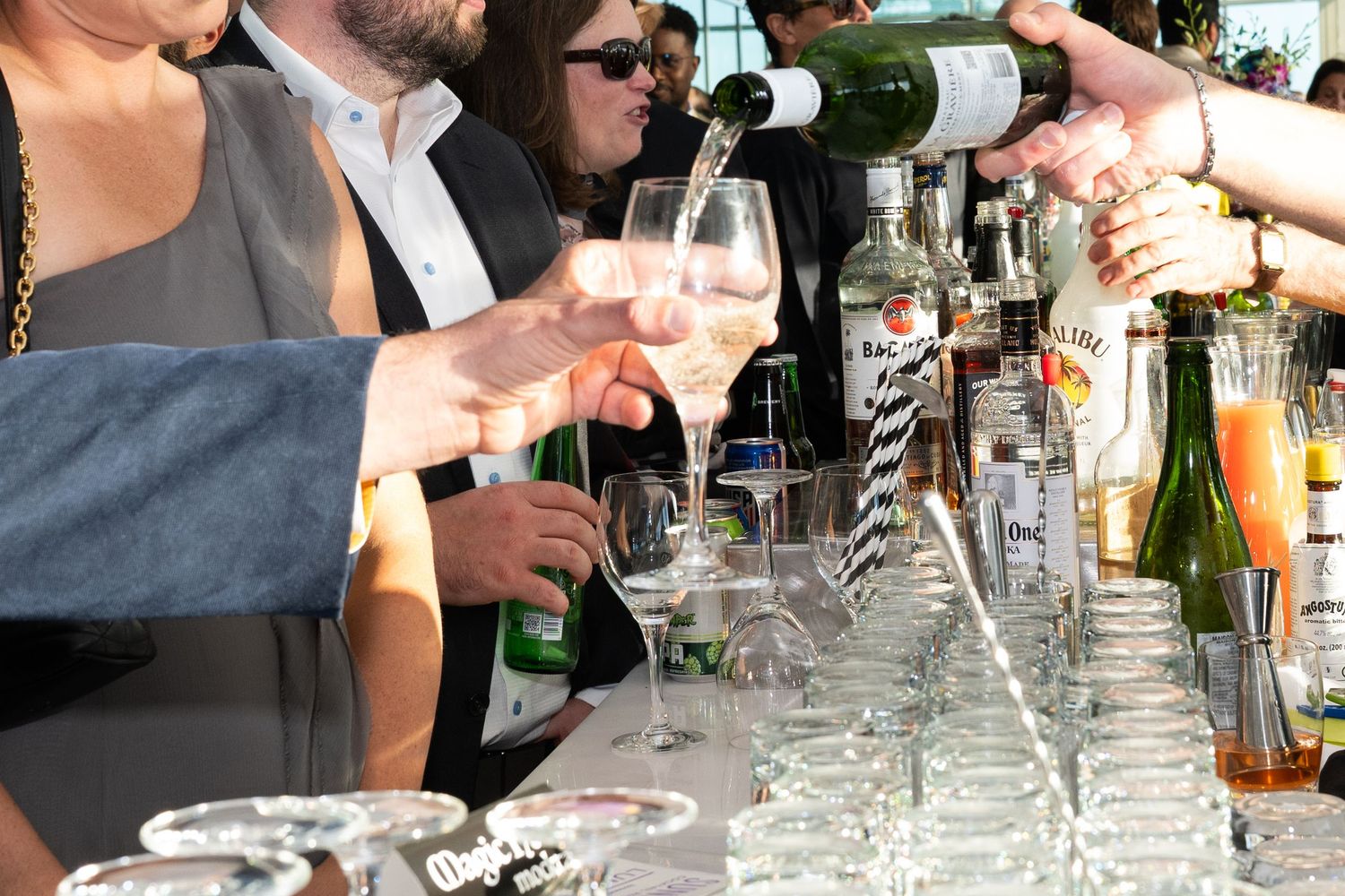 Guests holding wine glasses being filled with white wine at a busy outdoor bar with bottles on display.