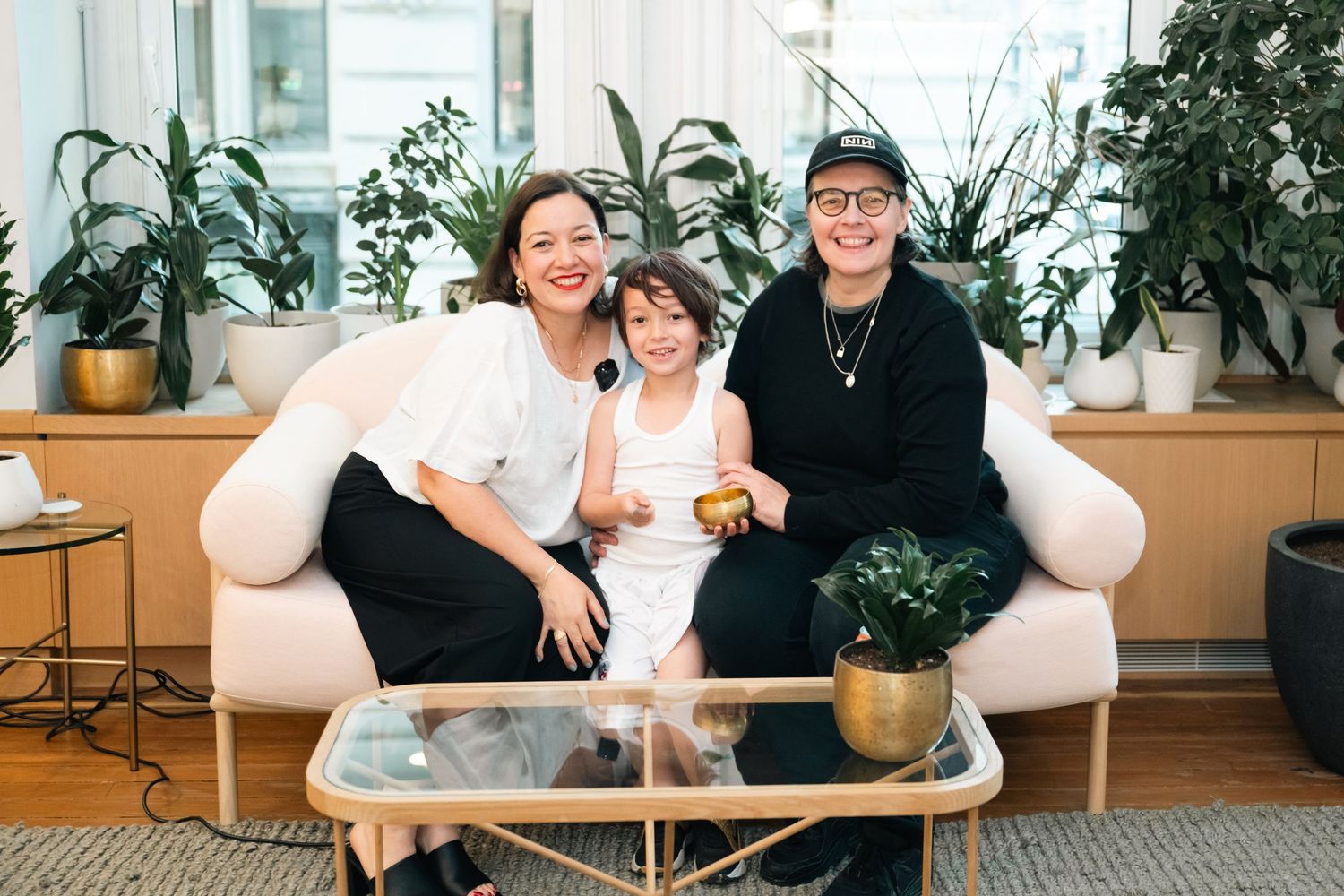 Two women and a child smile together on a white sofa surrounded by lush green plants in a bright modern space.
