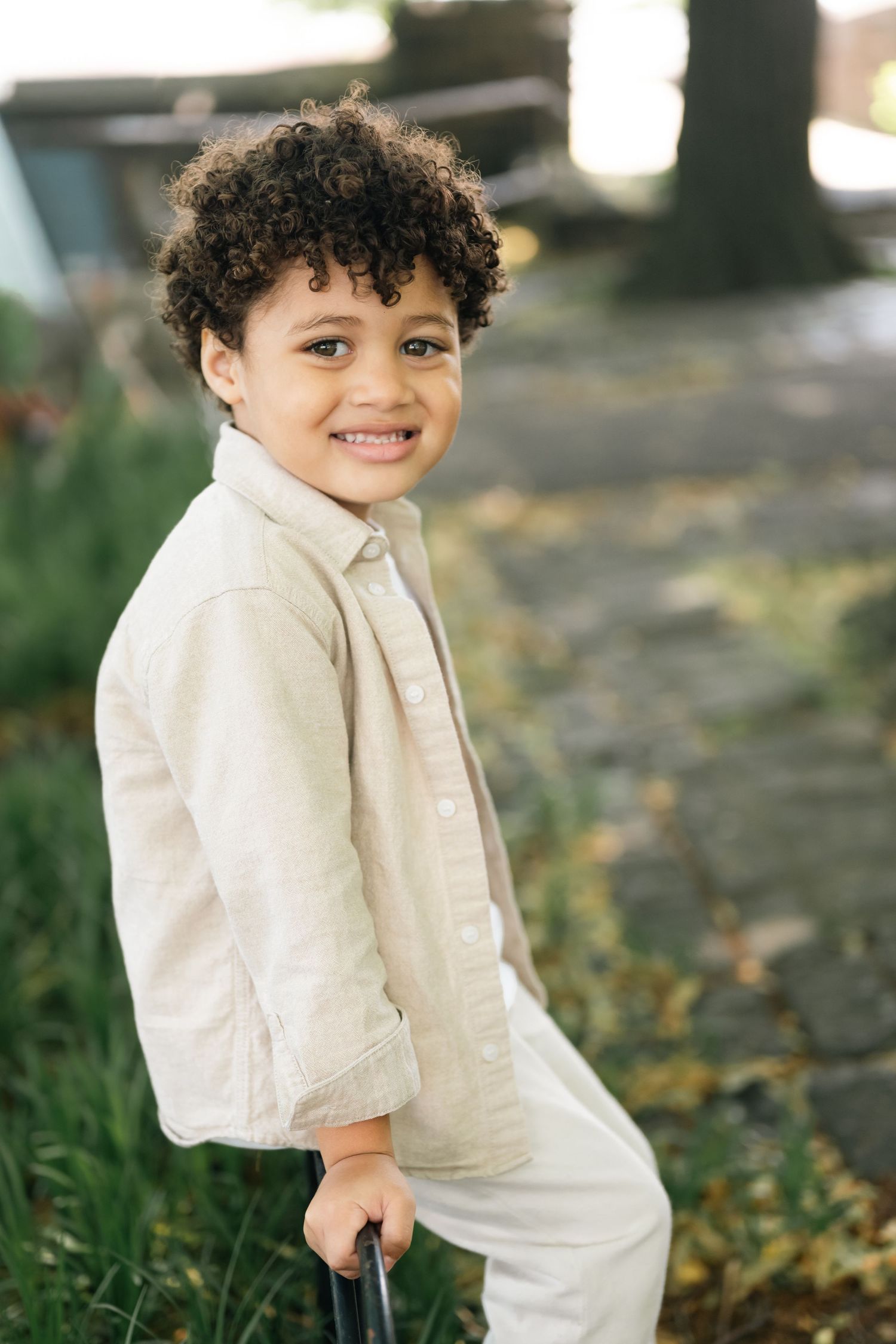 Smiling young boy with curly hair wearing a beige linen outfit, posing outdoors in a garden setting.