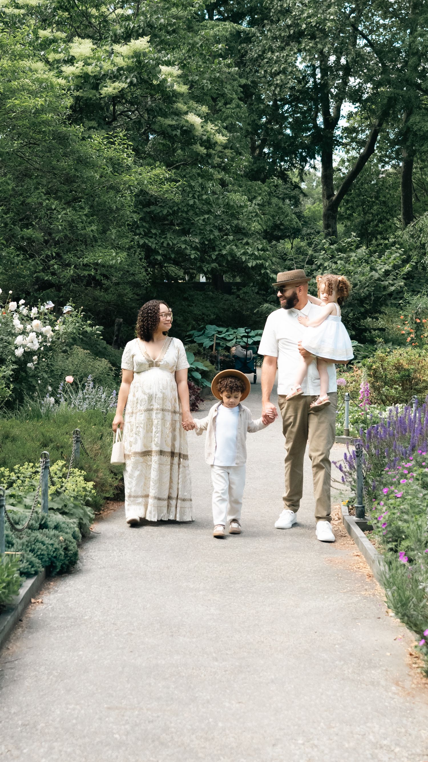 A family of four walks through a lush garden path, with a pregnant mother, father carrying a child, and a toddler in a hat.