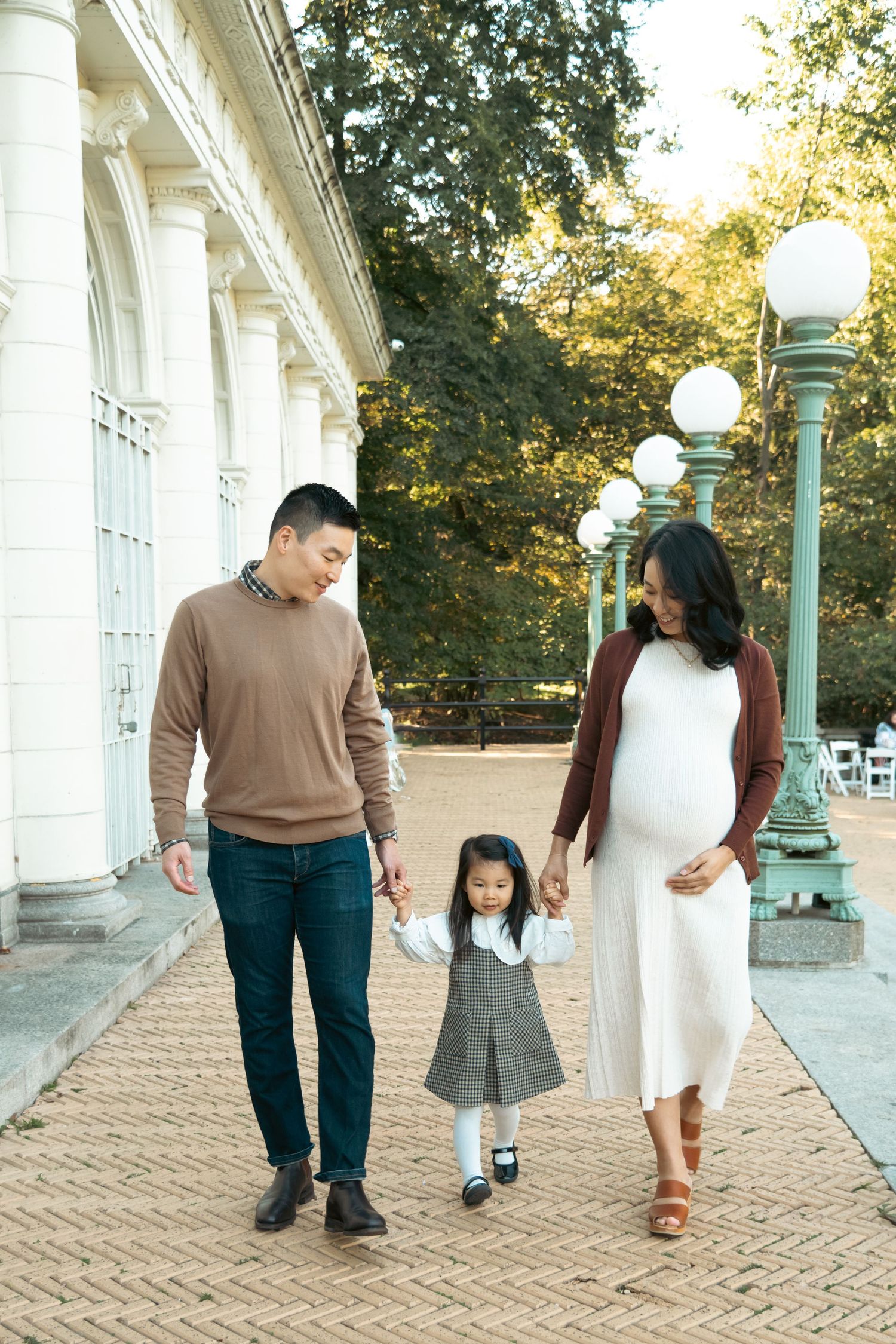 Pregnant Asian woman walks with husband and toddler daughter outside elegant white building in autumn park.