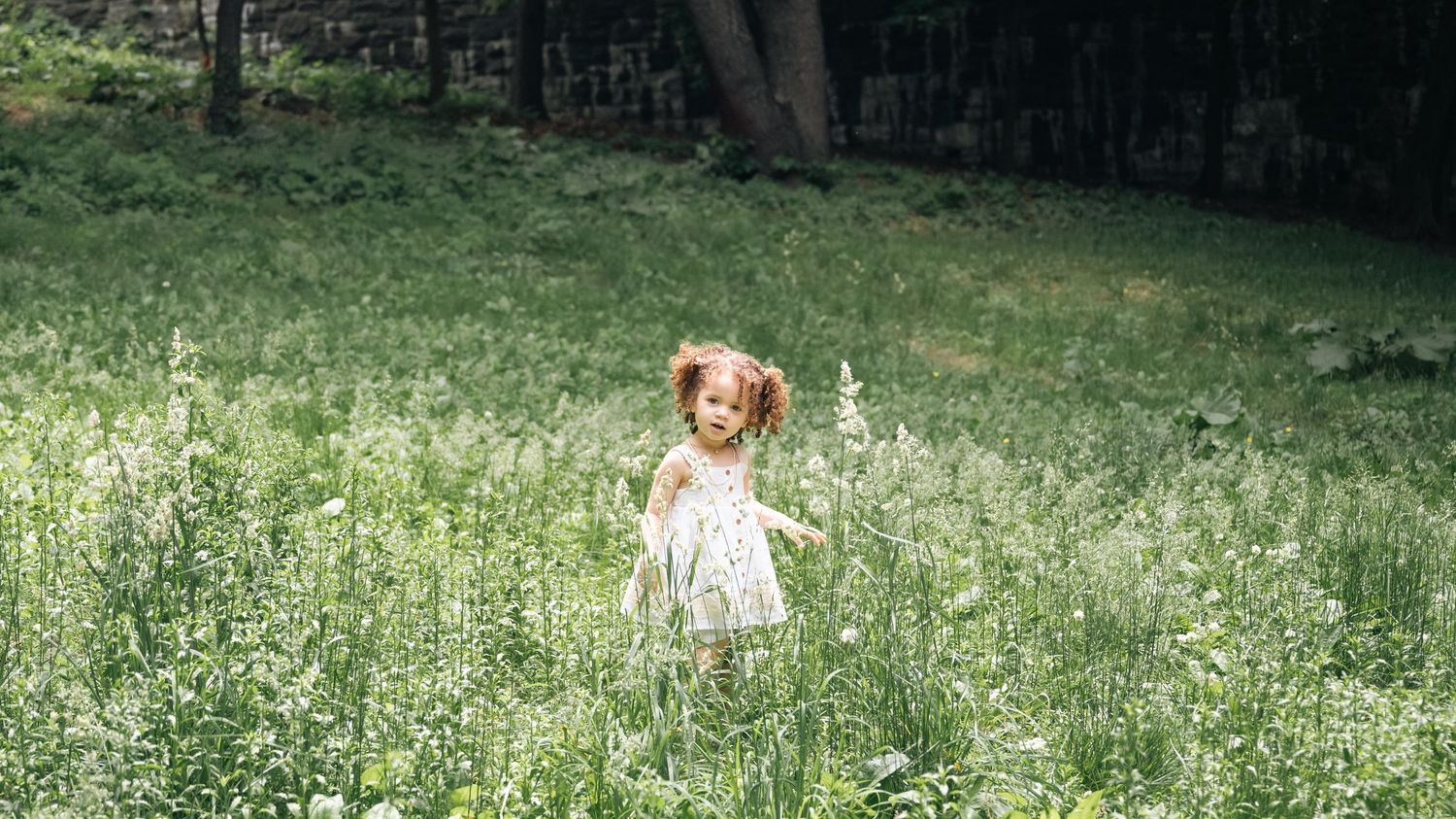 A young girl with red pigtails in a white dress runs through a lush green wildflower meadow near a forest.