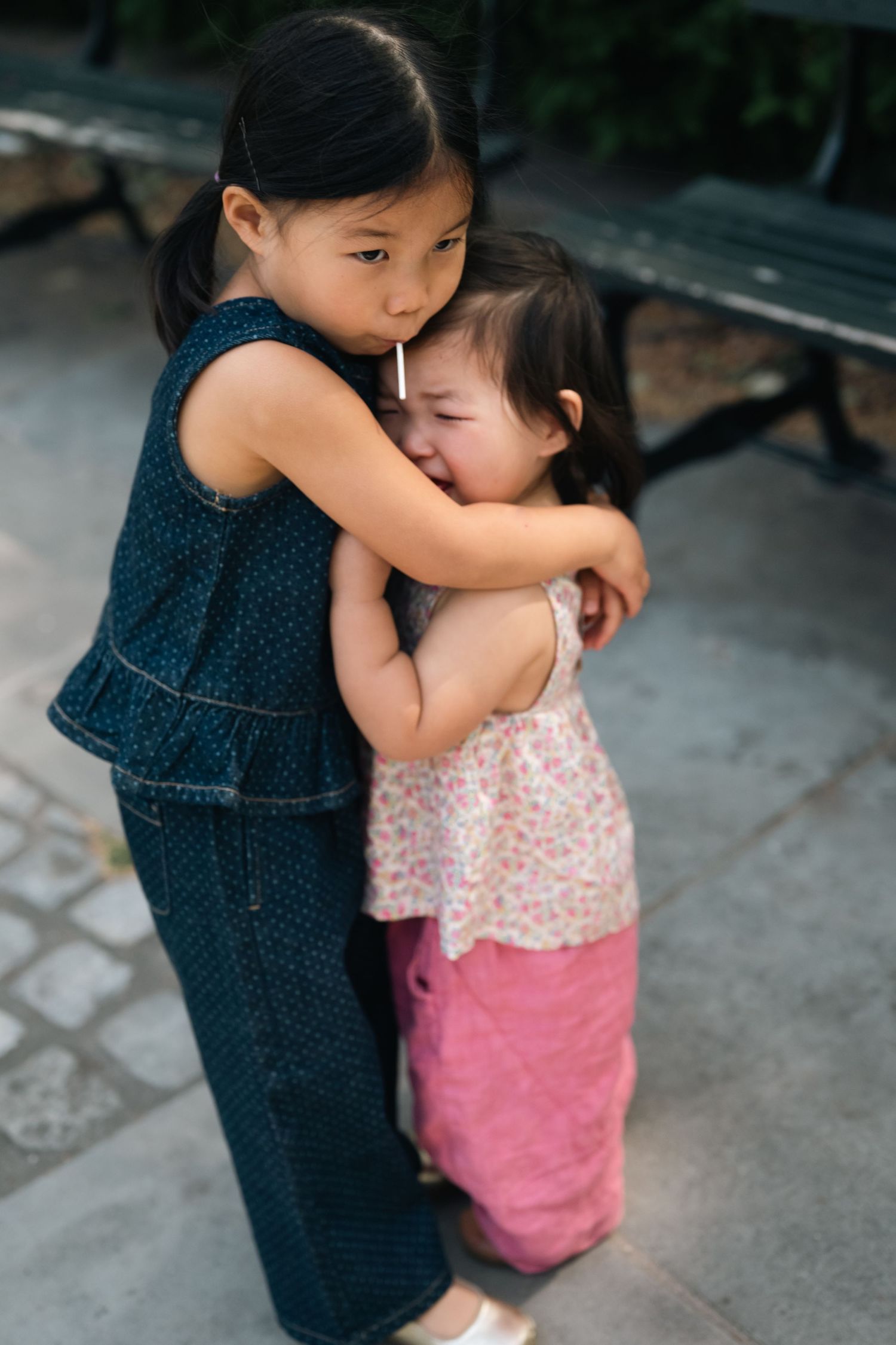 Two young Asian sisters share a sweet hug outdoors, with the older girl comforting her crying younger sibling.