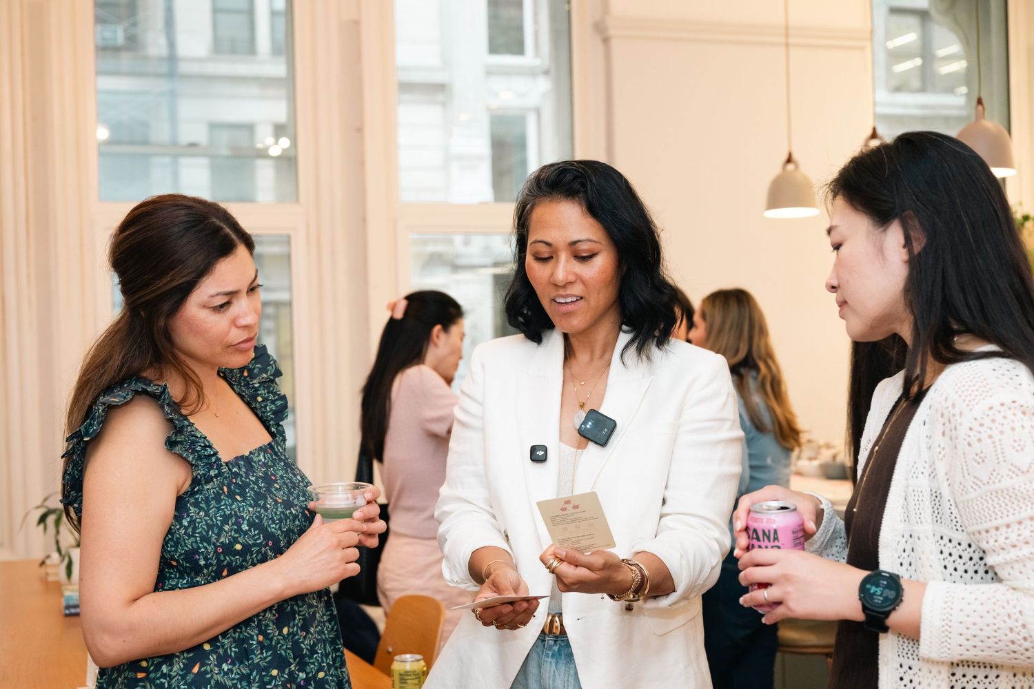 Three women networking at a social event, looking at a phone while holding drinks in a bright modern venue.