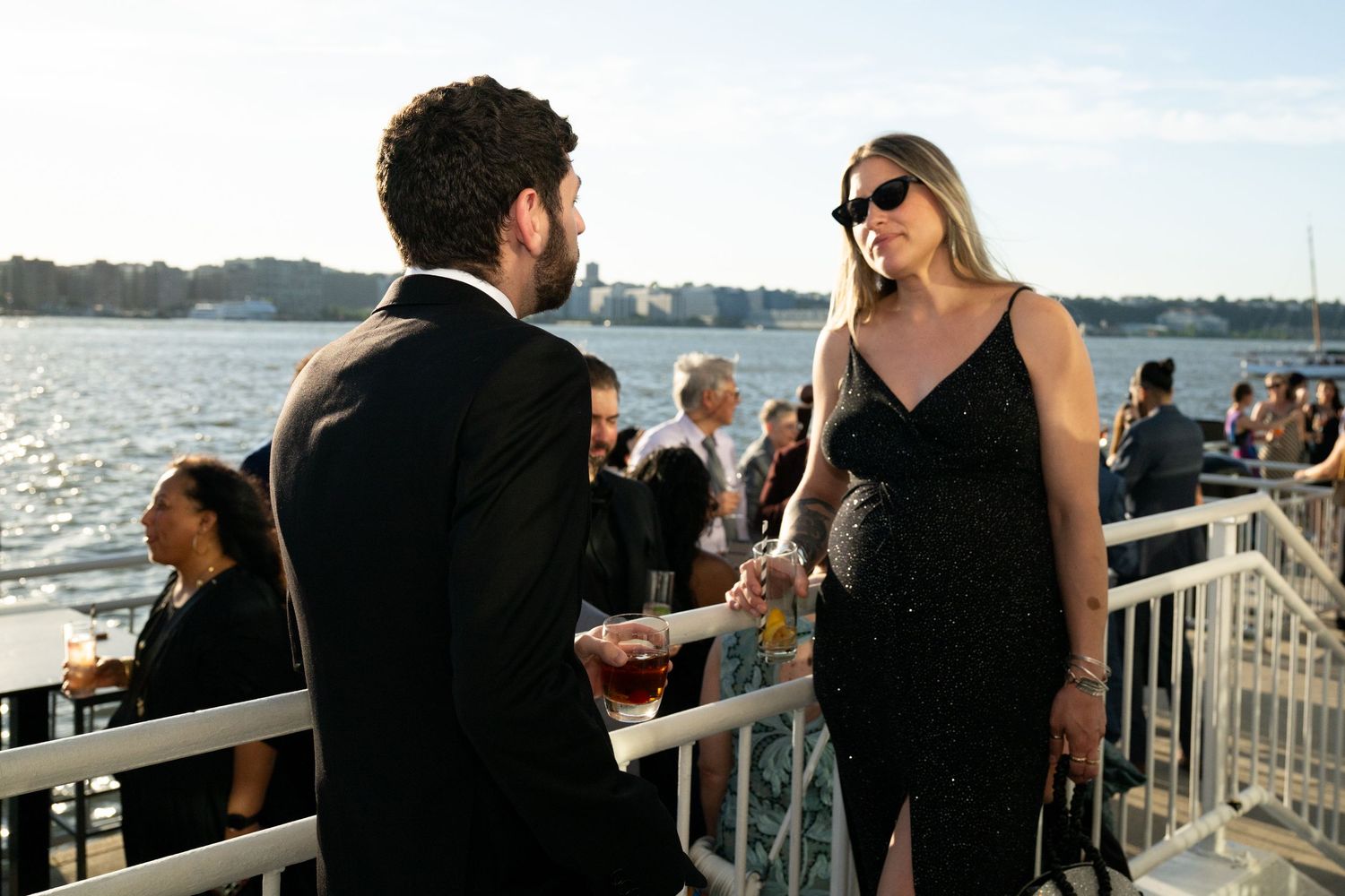 A couple in formal black attire converse on a waterfront terrace during an evening gala event.