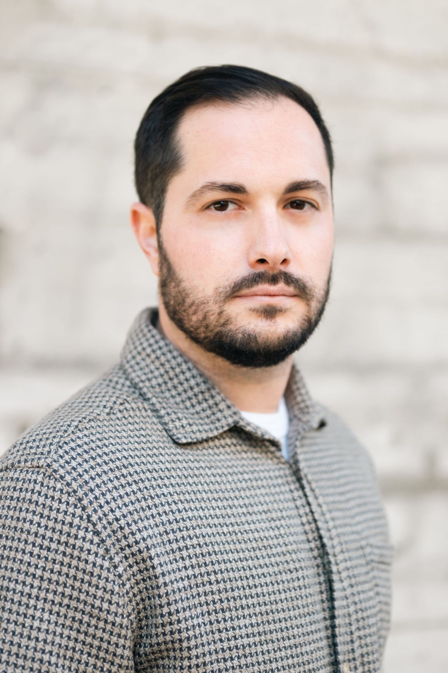 Professional headshot of man in gray patterned jacket against light wall.