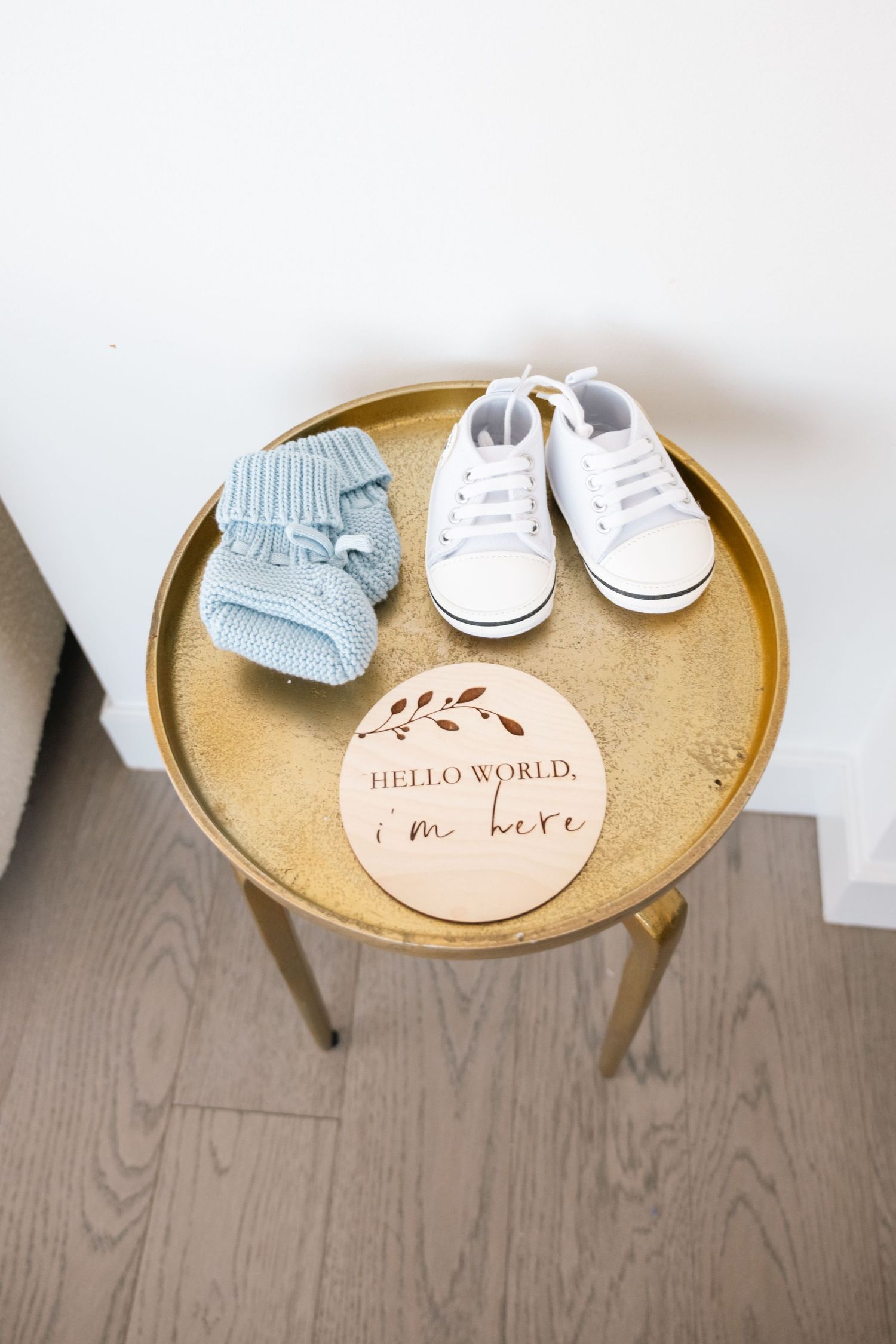 Baby shoes and booties displayed on gold side table with 'Hello World' welcome sign.