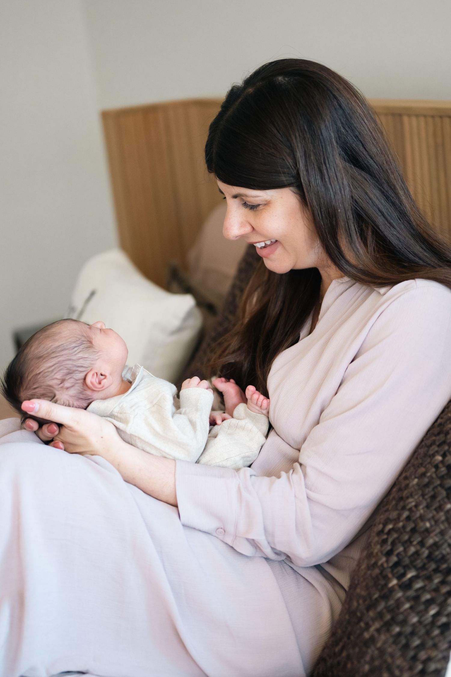 Parent in pink sweater holding sleeping newborn baby on couch in natural light setting.